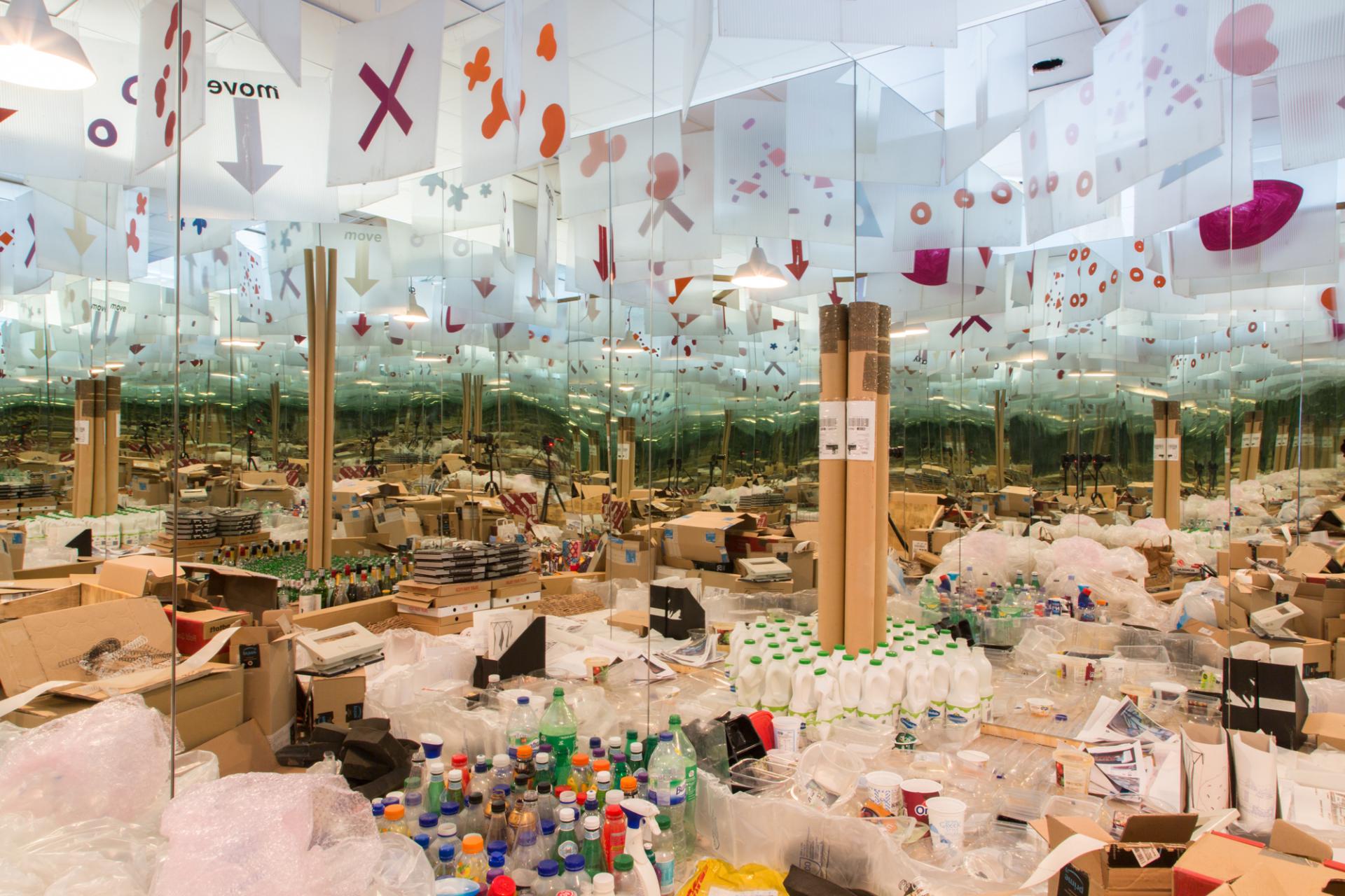 Art installation highlighting consumer waste, featuring mirrors and scattered plastic and cardboard materials, promoting sustainable design practices.