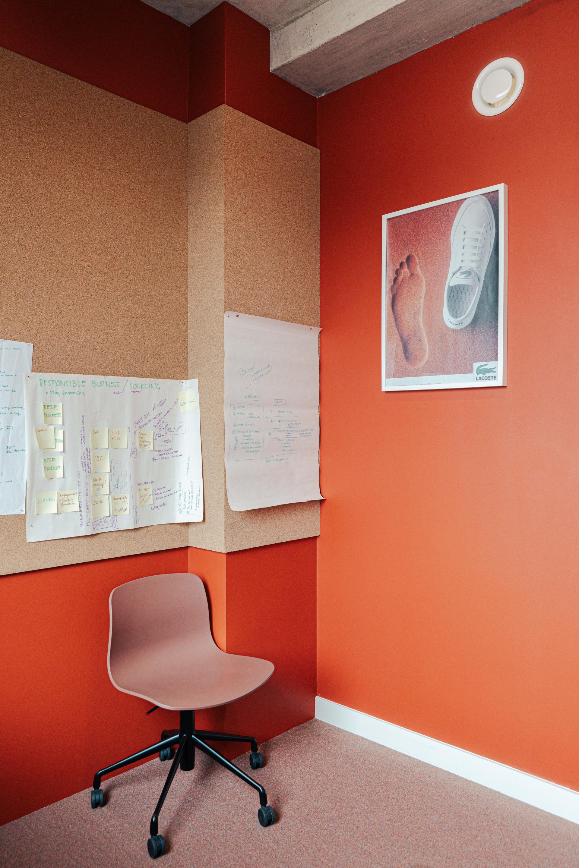 Modern workspace featuring a corkboard, planning notes, and a stylish chair against an orange wall, reflecting Lacoste’s design ethos.