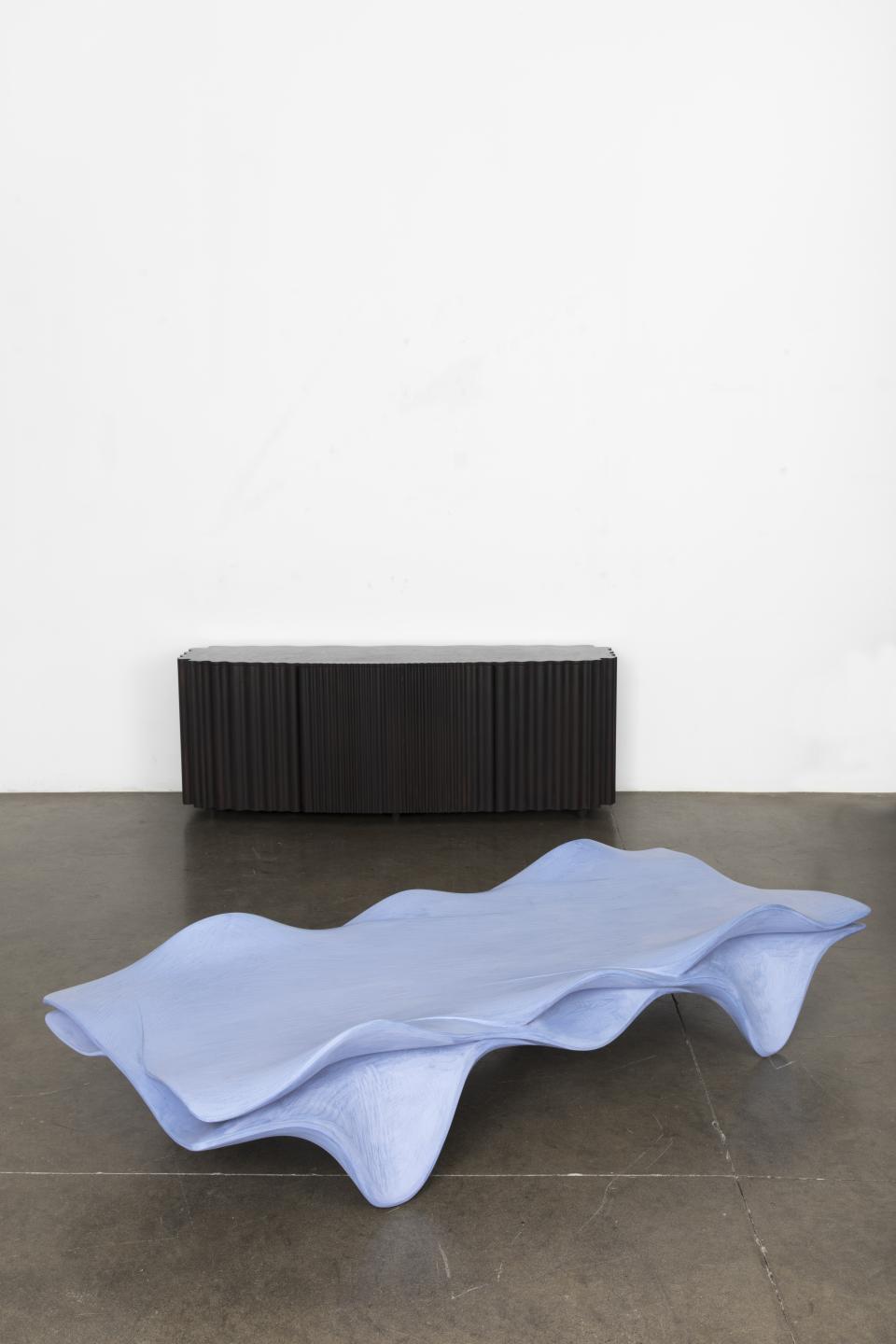 Sculptural blue coffee table with wave-like design, displayed in a minimalist gallery alongside a black, ribbed cabinet.