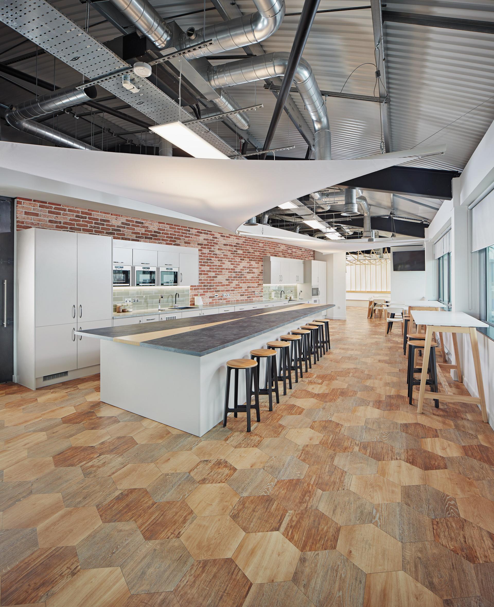 Modern workspace featuring a kitchen with a long counter, stylish stools, and hexagonal wooden flooring, embodying workplace-hospitality design.