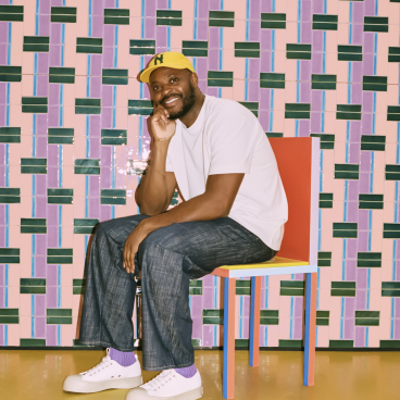 Yinka Ilori poses on a colorful chair against a vibrant, patterned tile wall at Clerkenwell Design Week.