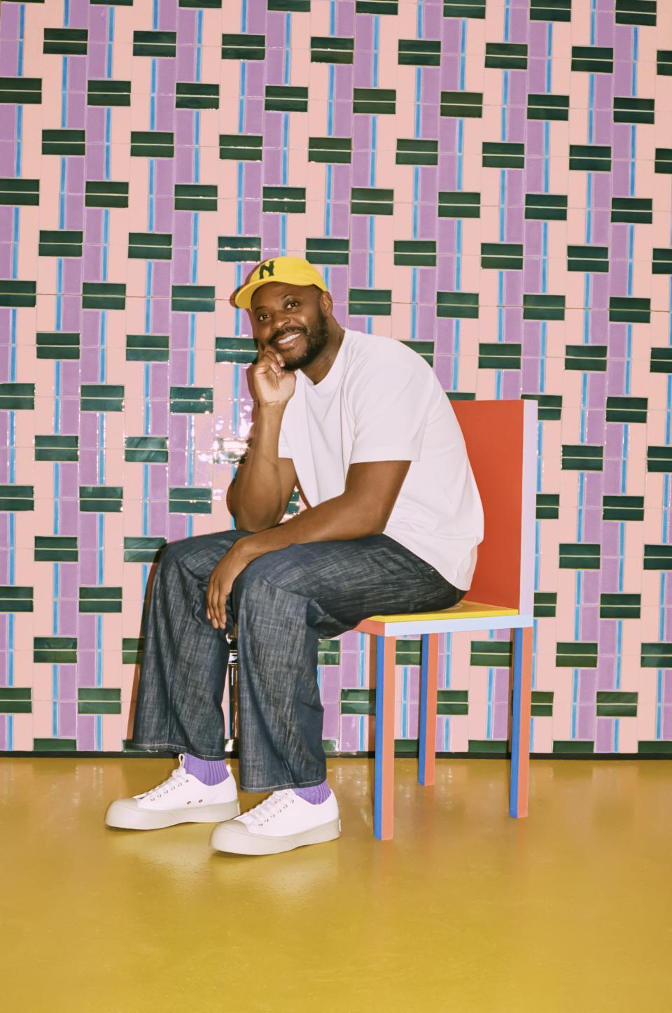 Yinka Ilori poses on a colorful chair against a vibrant, patterned tile wall at Clerkenwell Design Week.