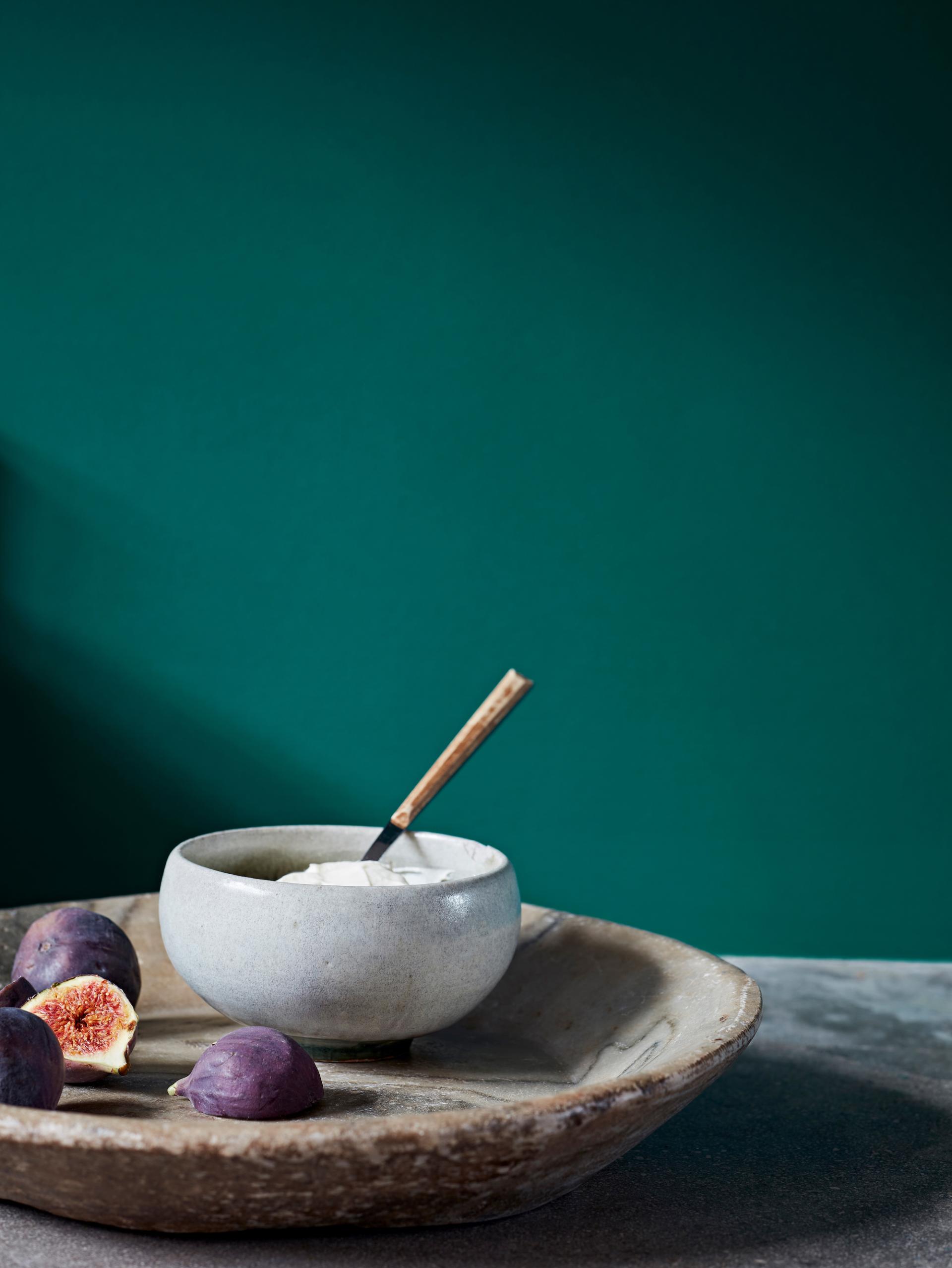 A minimalist arrangement featuring a gray bowl with a spoon, fresh figs, and a deep green wall backdrop.