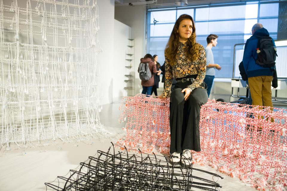 A woman sits on a colorful woven design piece at Dutch Design Week, surrounded by innovative installations.