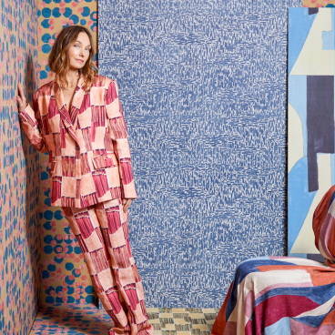 Interior designer Kelly Wearstler poses in a vibrant, patterned suit against colorful wallpaper and fabric backgrounds.