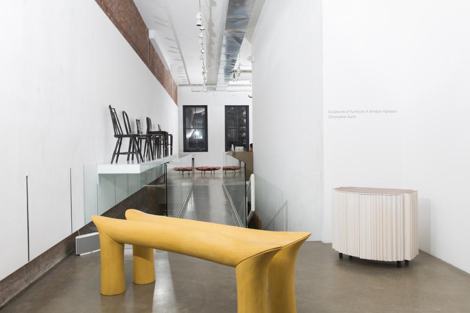 Sculptural furniture by Christopher Kurtz, featuring a yellow table and a curved white cabinet in a gallery setting.