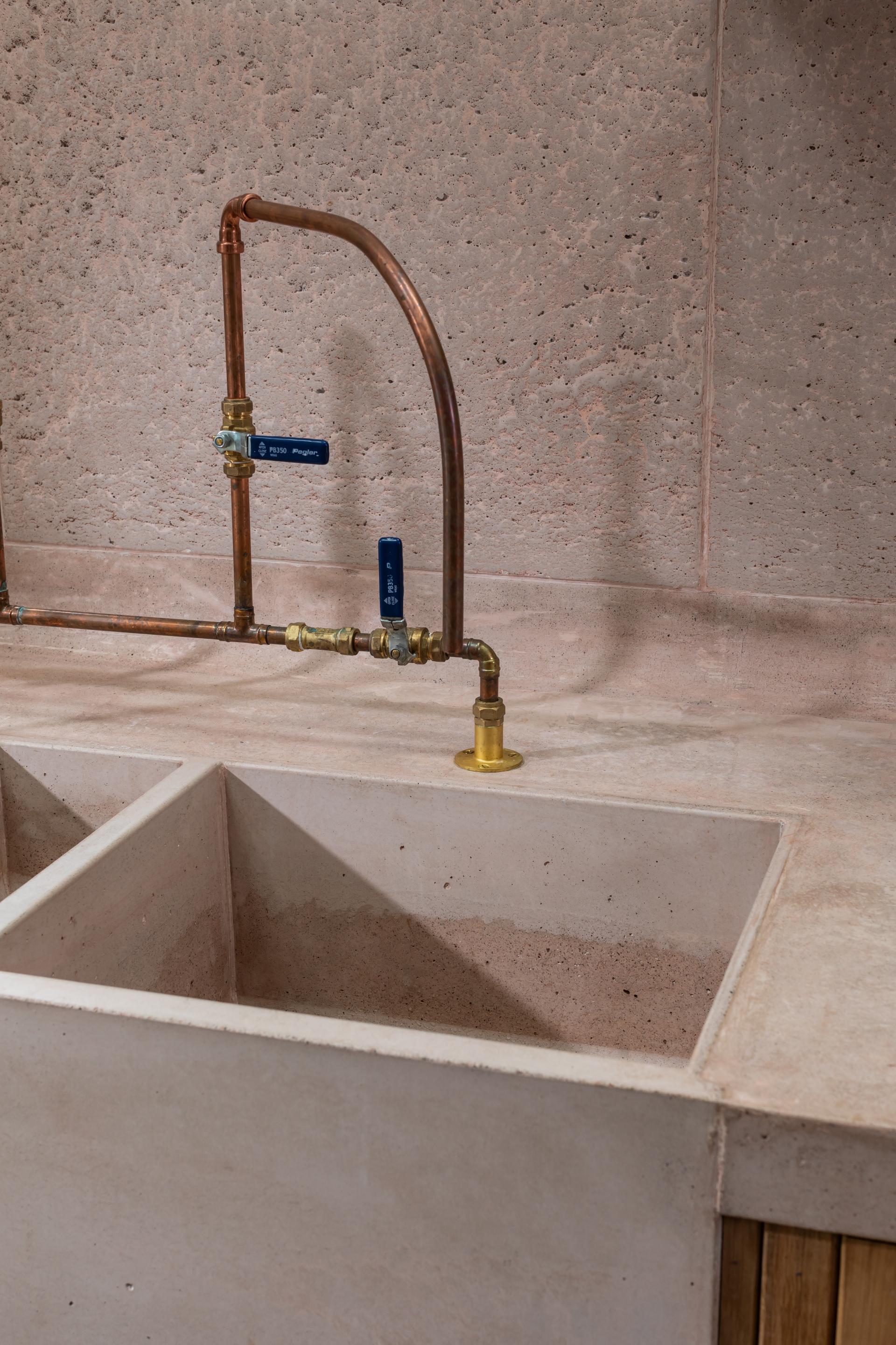 Poured pink concrete double sink with brass fittings and textured wall, showcasing modern kitchen design by Nicholas Denney Studio.