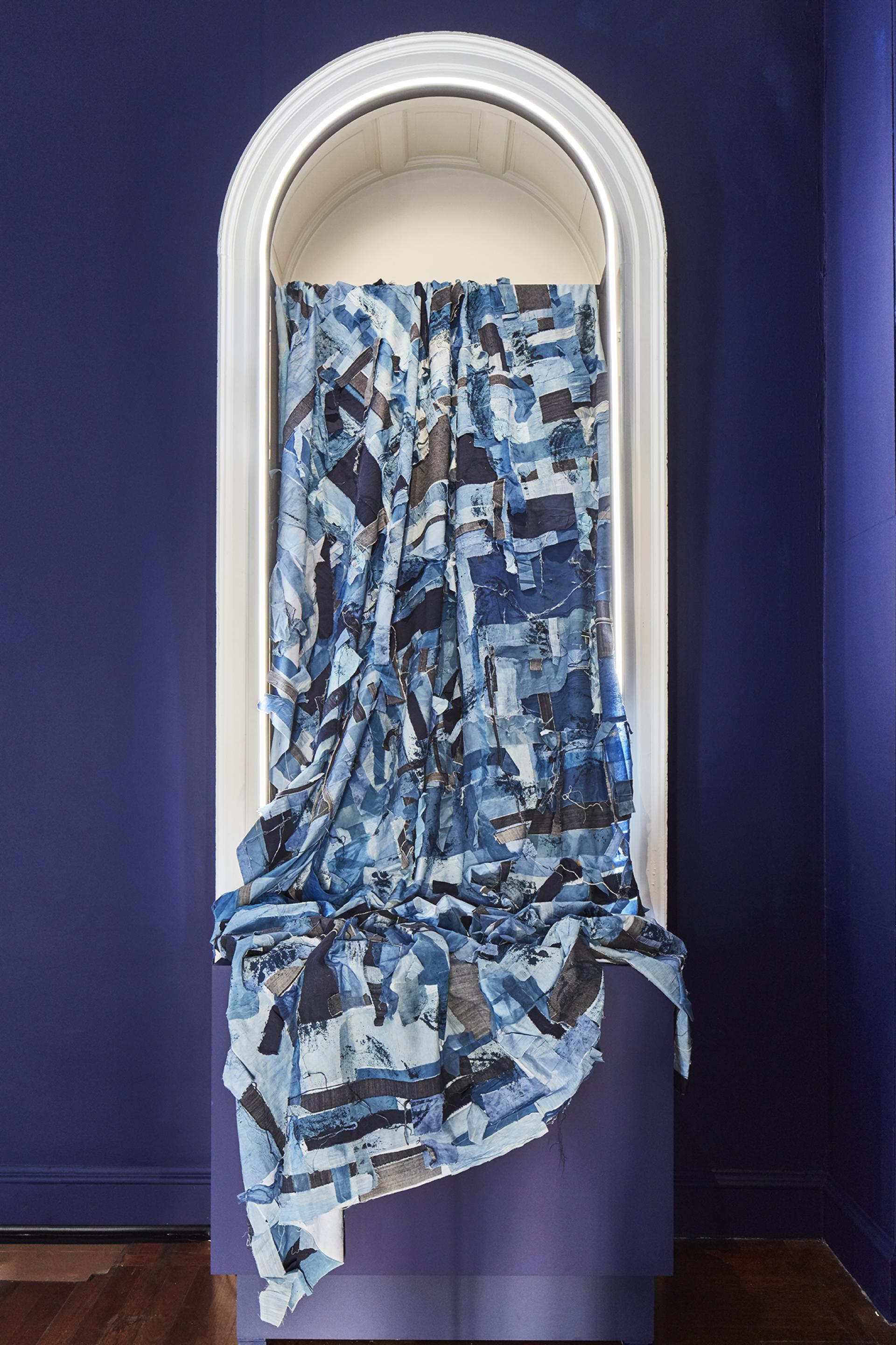 Textured blue fabric drapes elegantly in an arched alcove at the London Design Biennale, showcasing contemporary design.