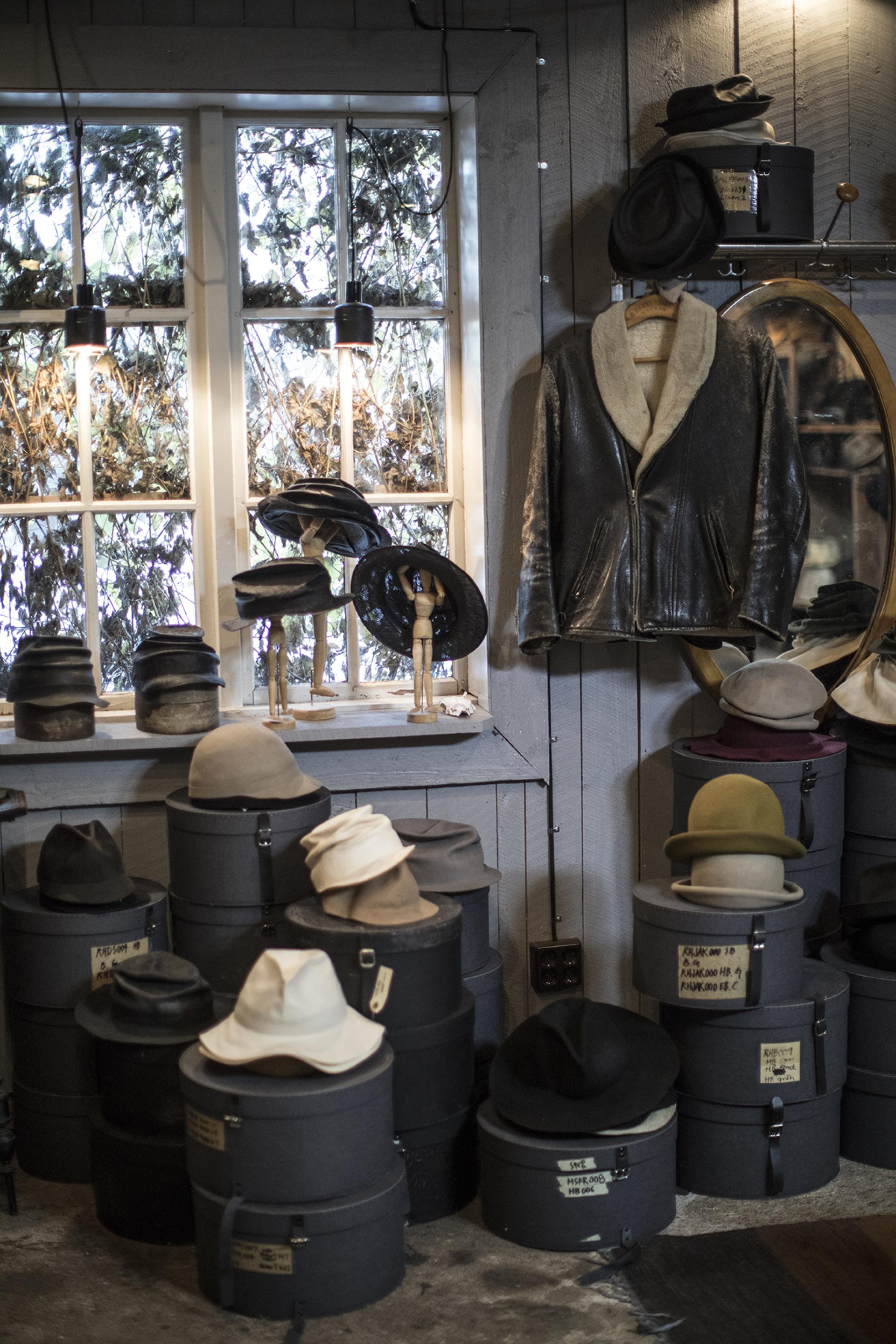 Inside a quaint workshop, various handcrafted hats are displayed atop storage boxes, surrounded by natural light and rustic decor.