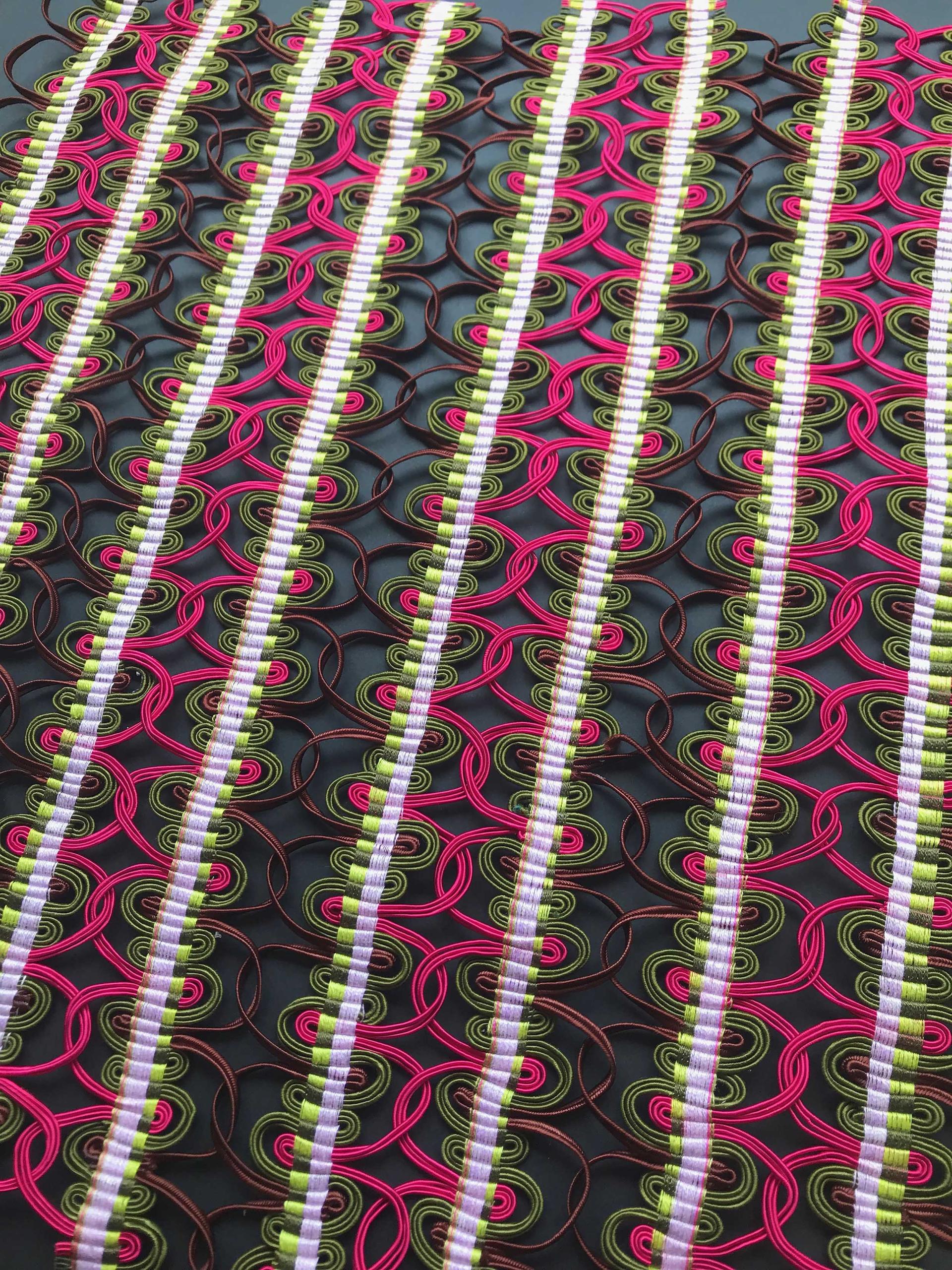 Vibrant hand-woven passementerie artwork featuring intricate patterns in pink, green, and brown threads on a dark background.