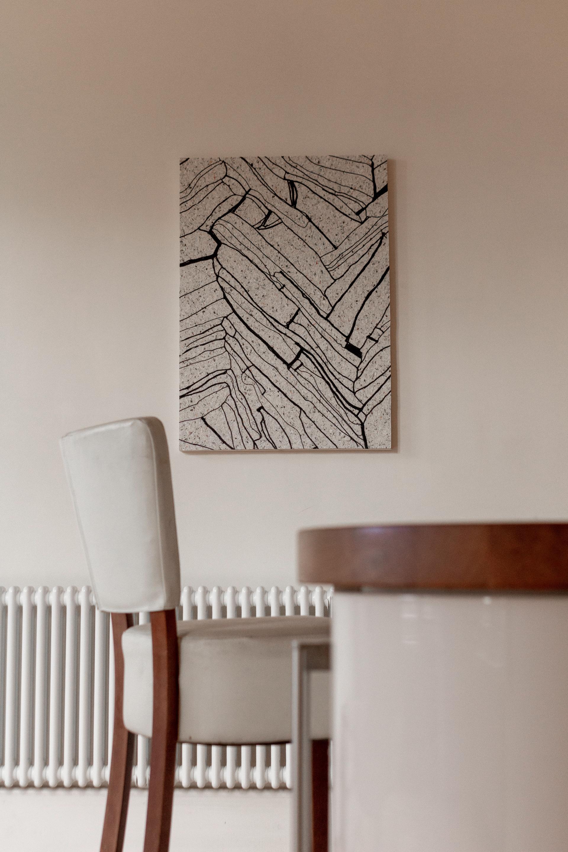 Textile art featuring abstract black lines on a speckled textured background, displayed above a modern chair and table.