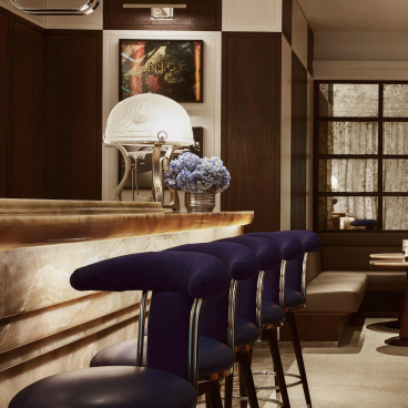Elegant bar scene with plush navy stools, modern decor, and a floral centerpiece at The Maybourne Bar.