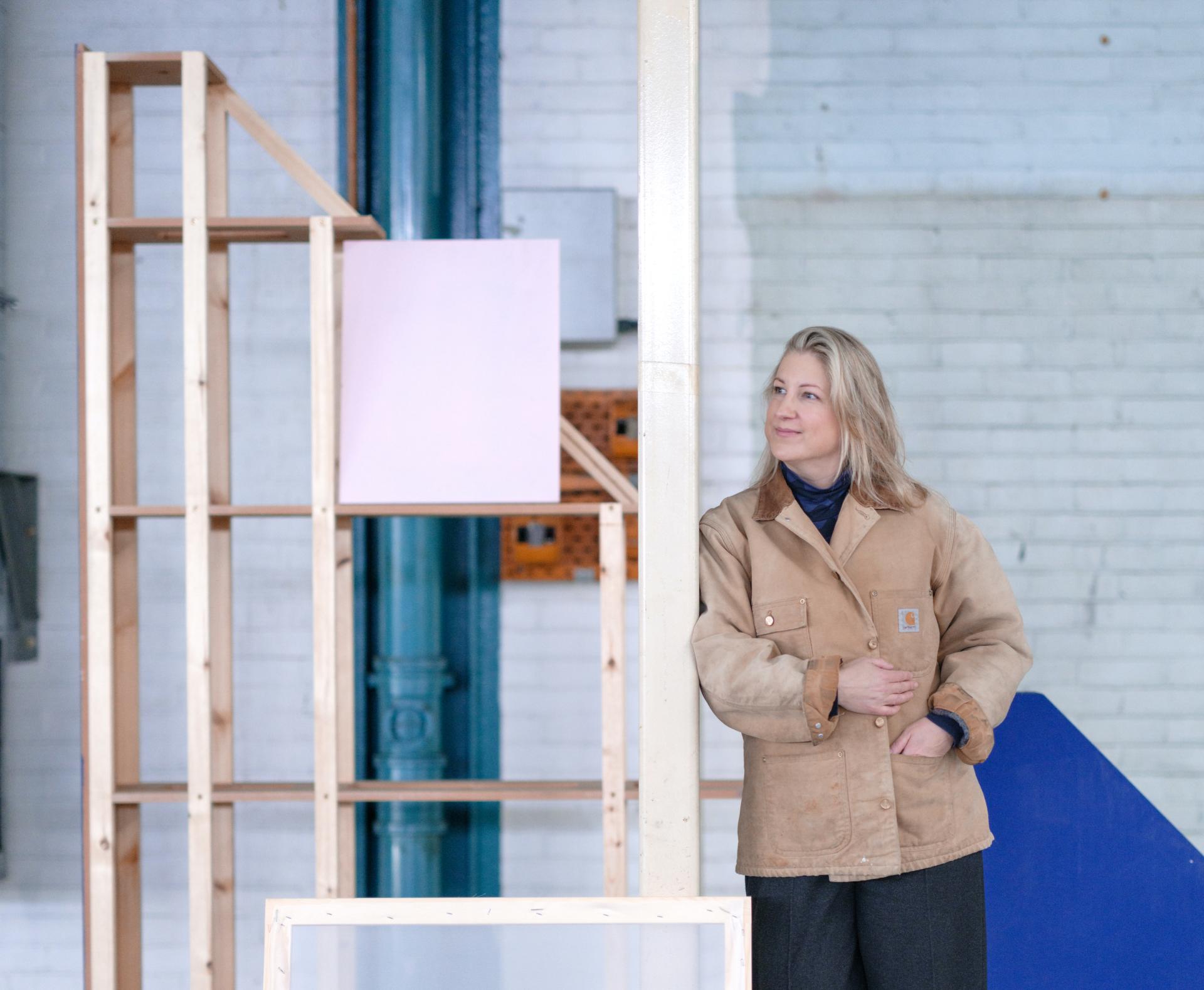 A woman in a brown coat stands beside a wooden exhibition structure at Dundee Design Festival 2024.