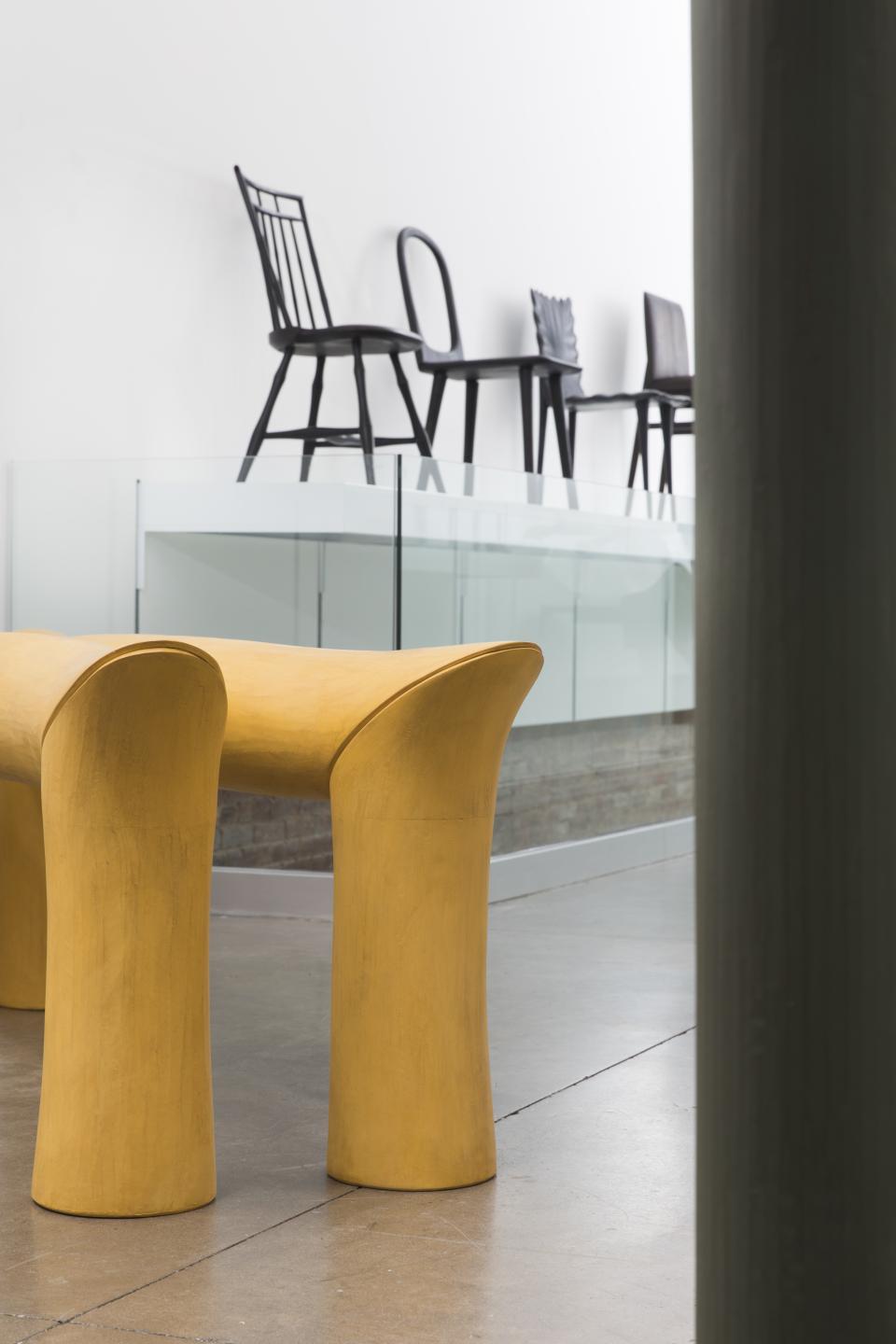 A yellow sculptural bench in the foreground with traditional Windsor chairs displayed in the background.