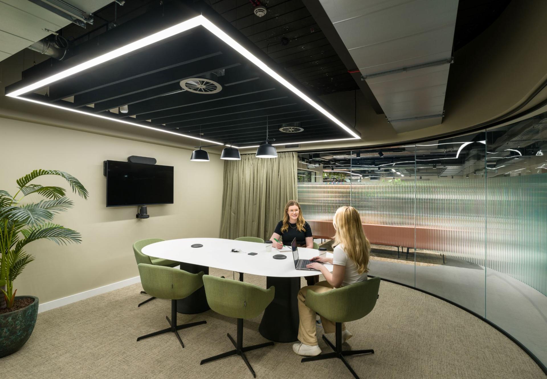 Modern meeting room at Glasshouse featuring green chairs, a large table, and collaborative workspace with women discussing ideas.