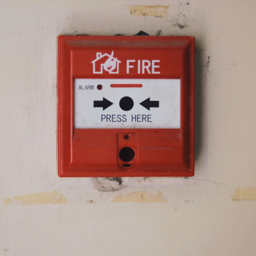 Red fire alarm panel mounted on a wall, featuring a "PRESS HERE" button for emergency notifications.