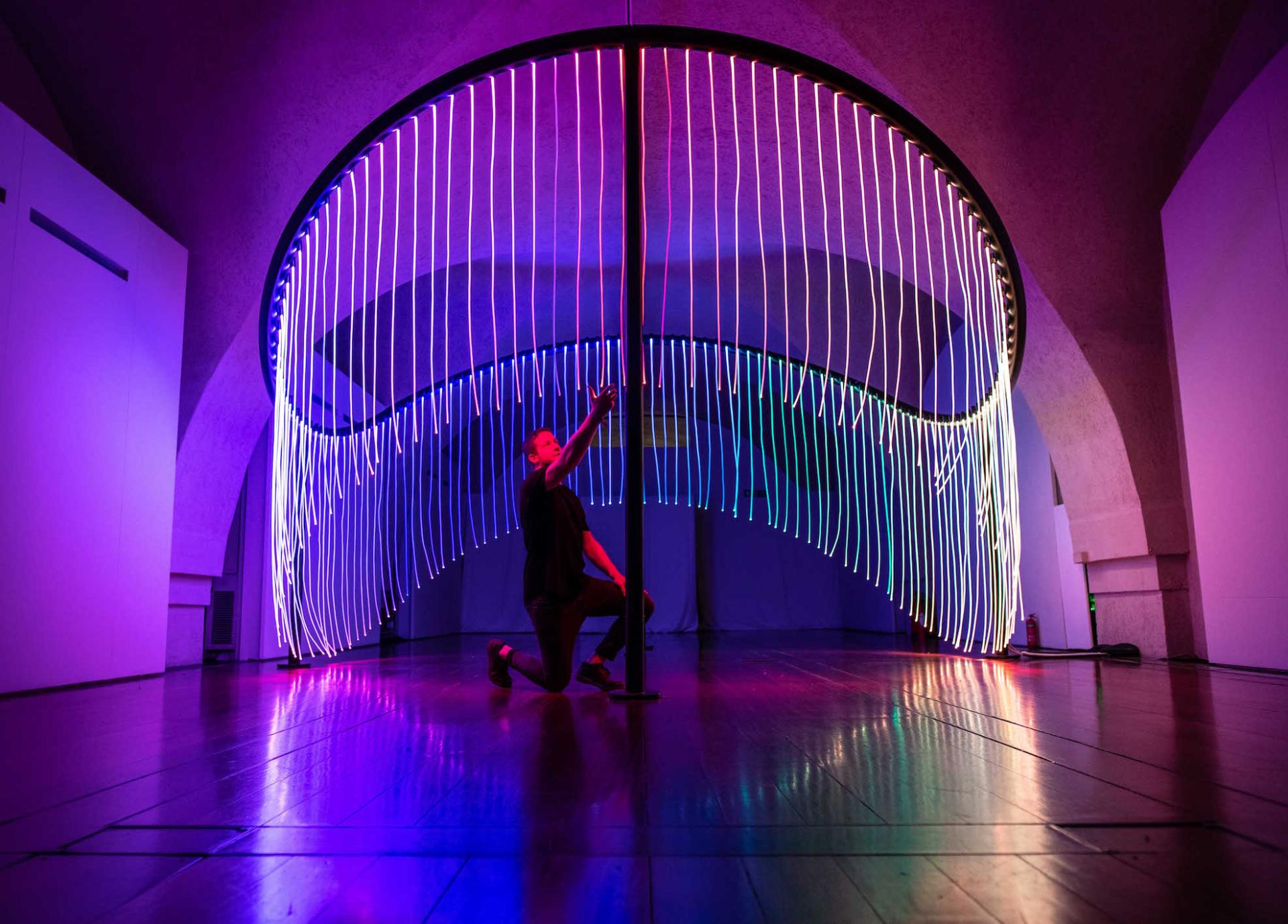 A performer interacts with vibrant, illuminated strands of light in a stylish exhibit at the London Design Biennale.