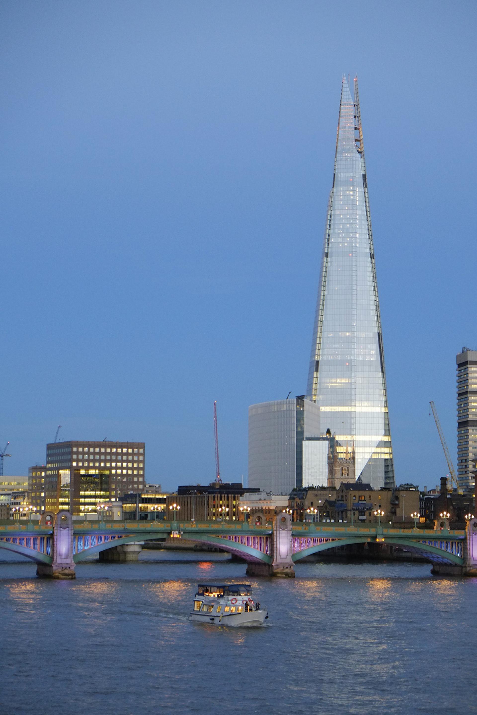 The Shard skyscraper rises above the Thames River, with a boat passing under an illuminated bridge in London.