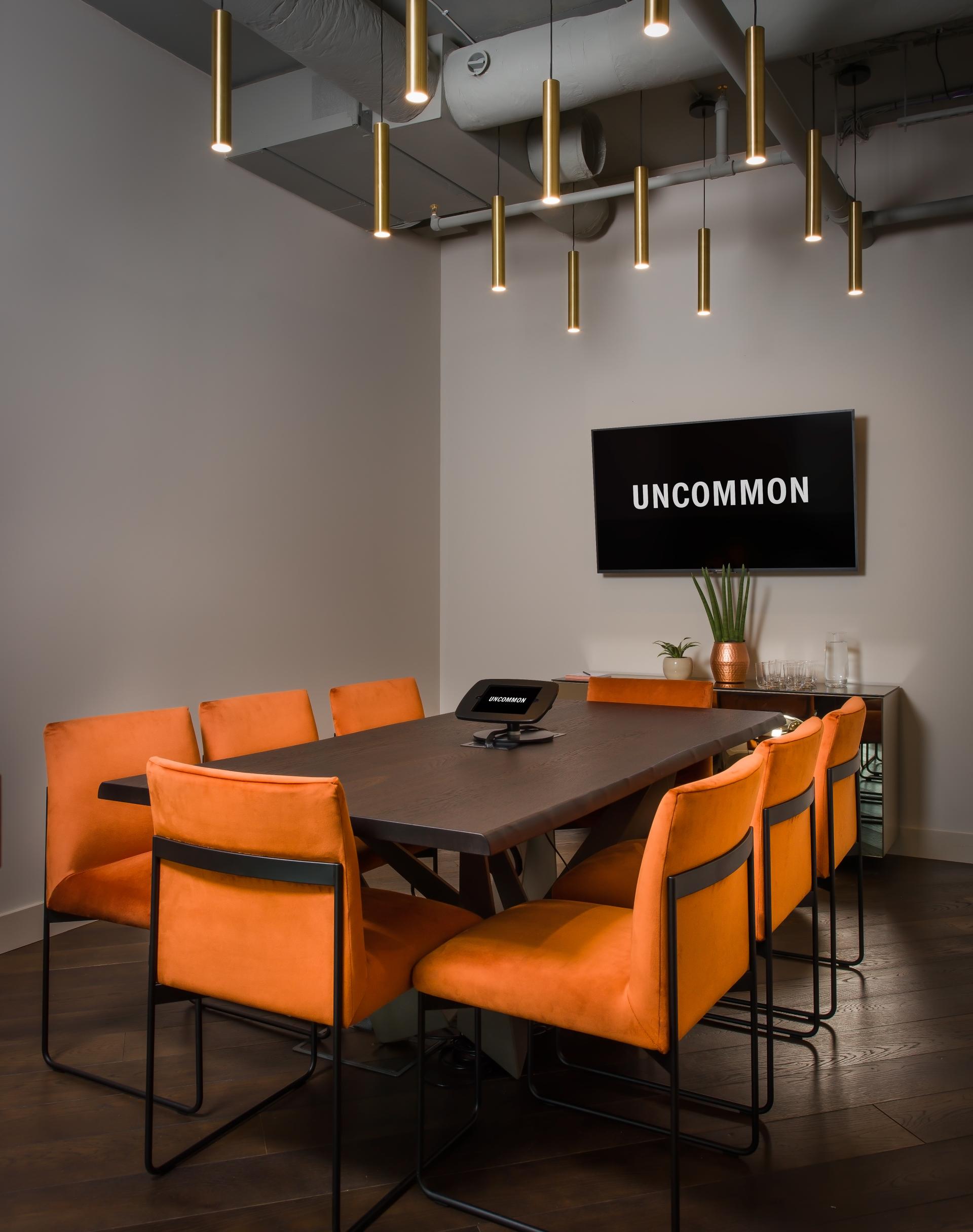 Modern conference room featuring a large table, orange chairs, pendant lighting, and a wall-mounted screen displaying "UNCOMMON."