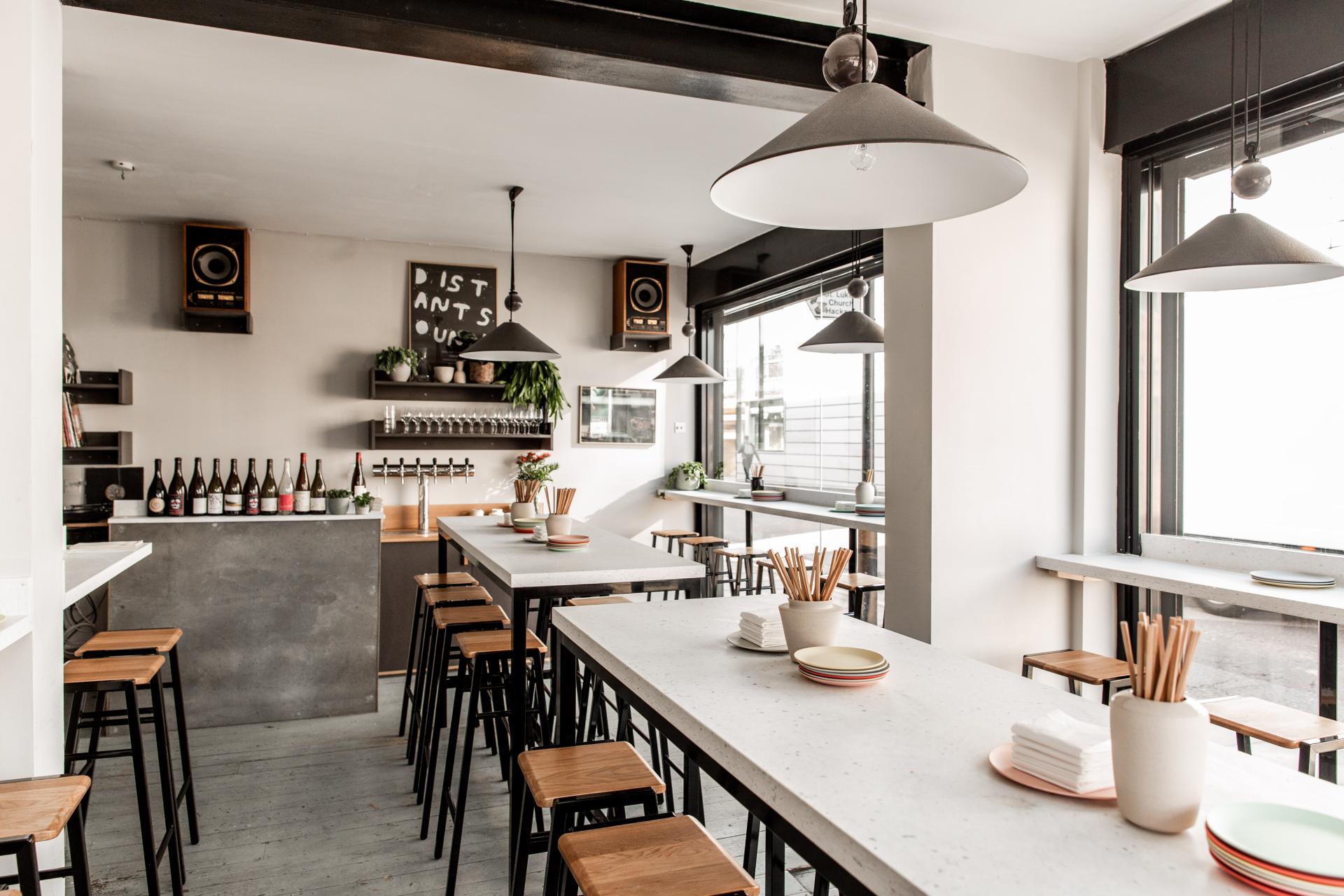 Modern restaurant interior featuring handcrafted tables, bar area, and minimalist decor with plants and natural light.