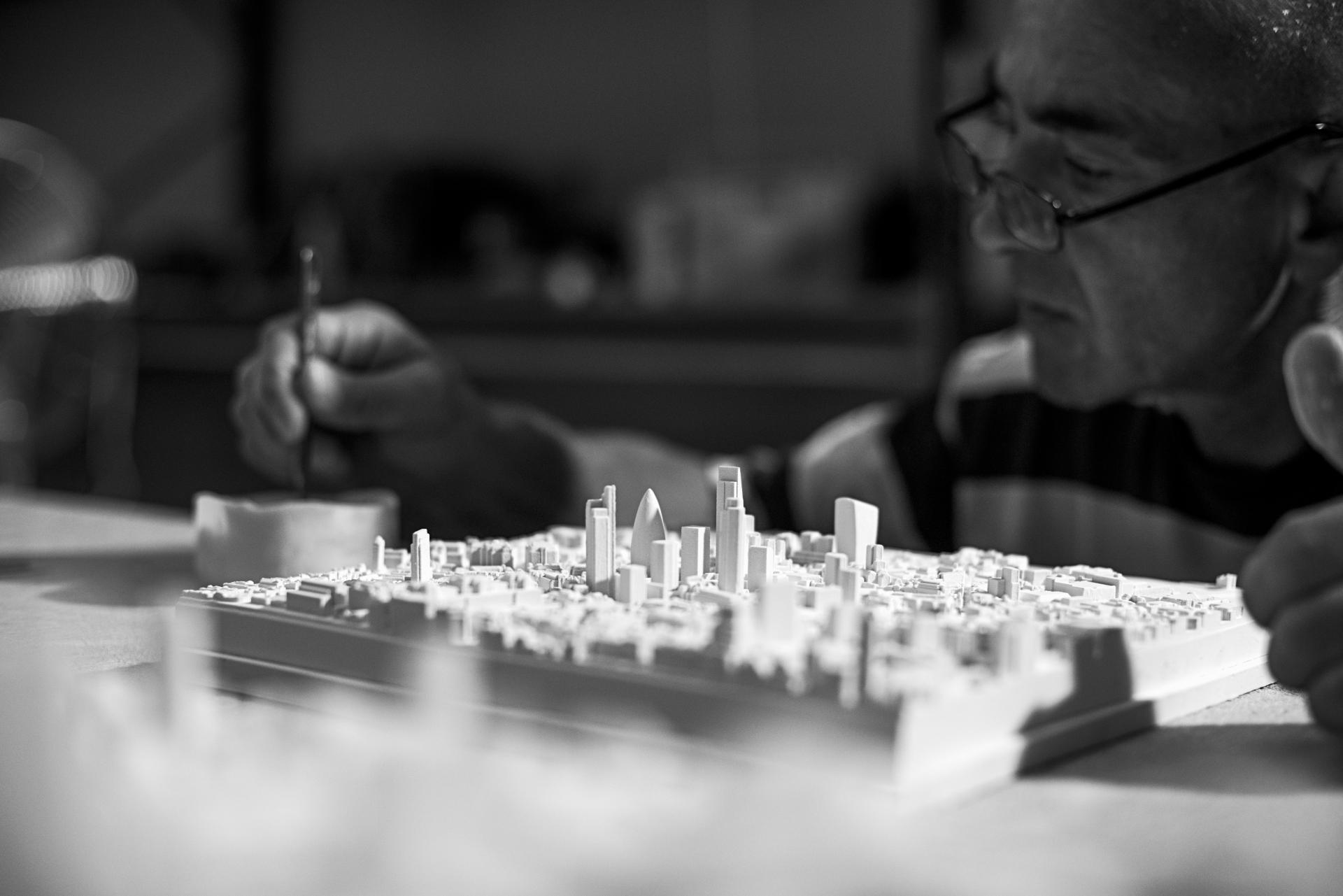 An artisan intricately details a three-dimensional scale model of an urban landscape, showcasing architectural features.