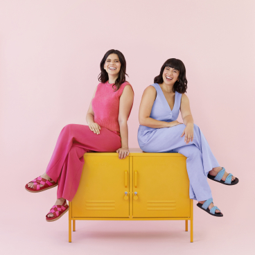 Becca Stern and Jess Gray, co-founders of Mustard Made, pose playfully on a bright yellow cabinet against a pink backdrop.