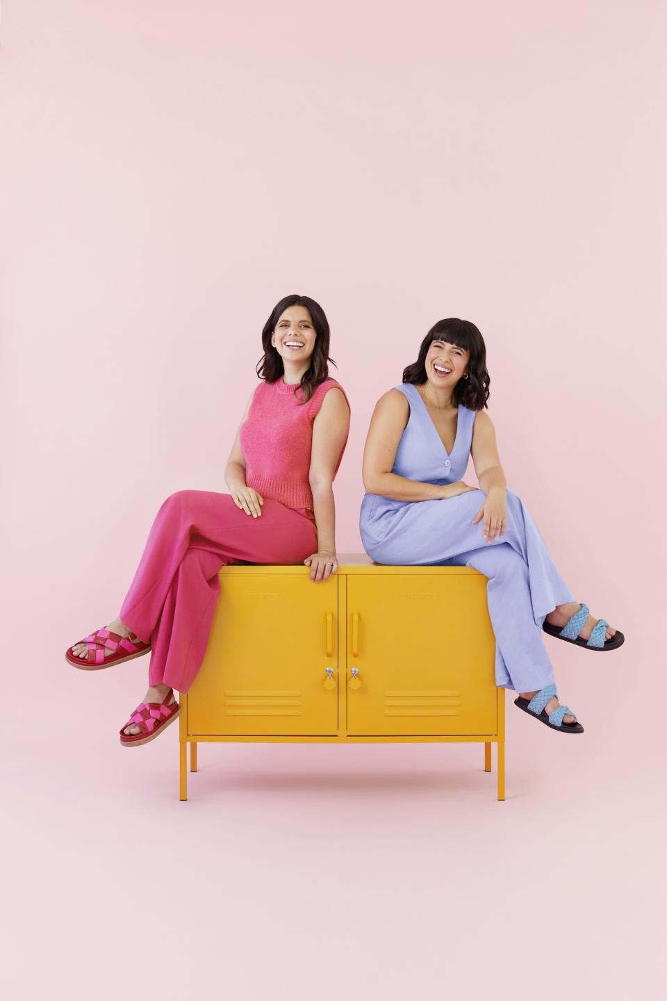 Becca Stern and Jess Gray, co-founders of Mustard Made, pose playfully on a bright yellow cabinet against a pink backdrop.