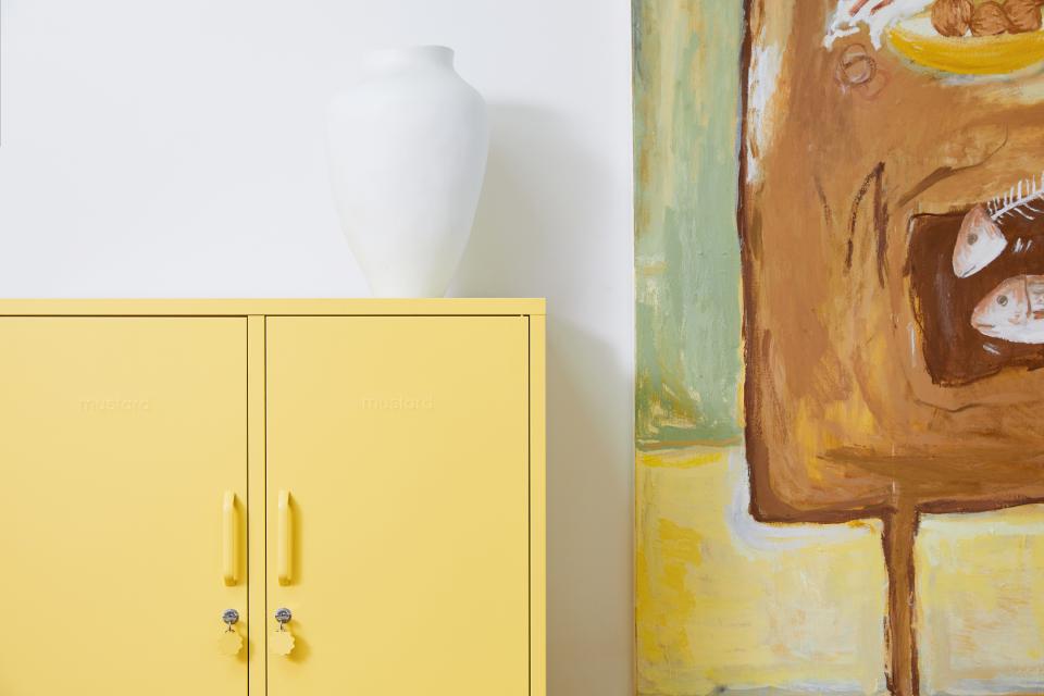Bright yellow Mustard Made cabinet with a white vase on top, beside a colorful abstract painting.