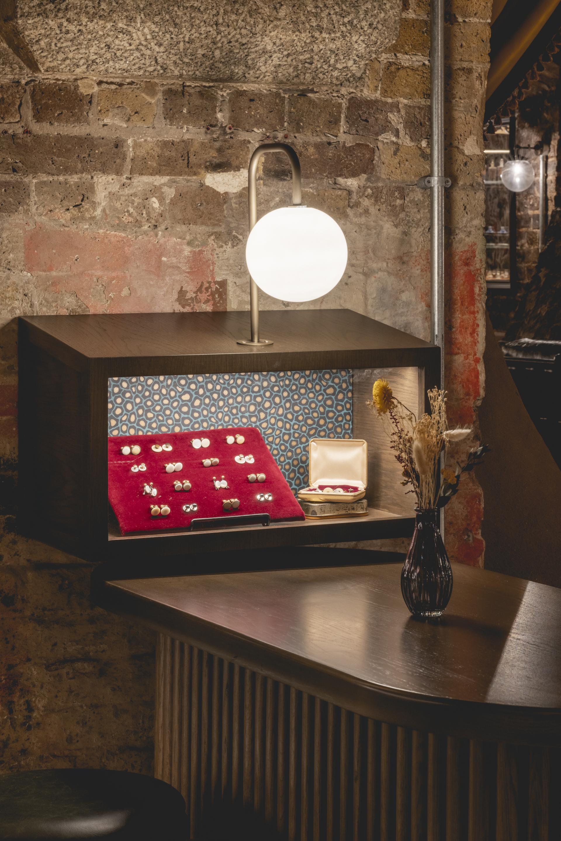 Elegant display case in The Libertine featuring jewelry, illuminated by modern lighting against a rustic brick wall.