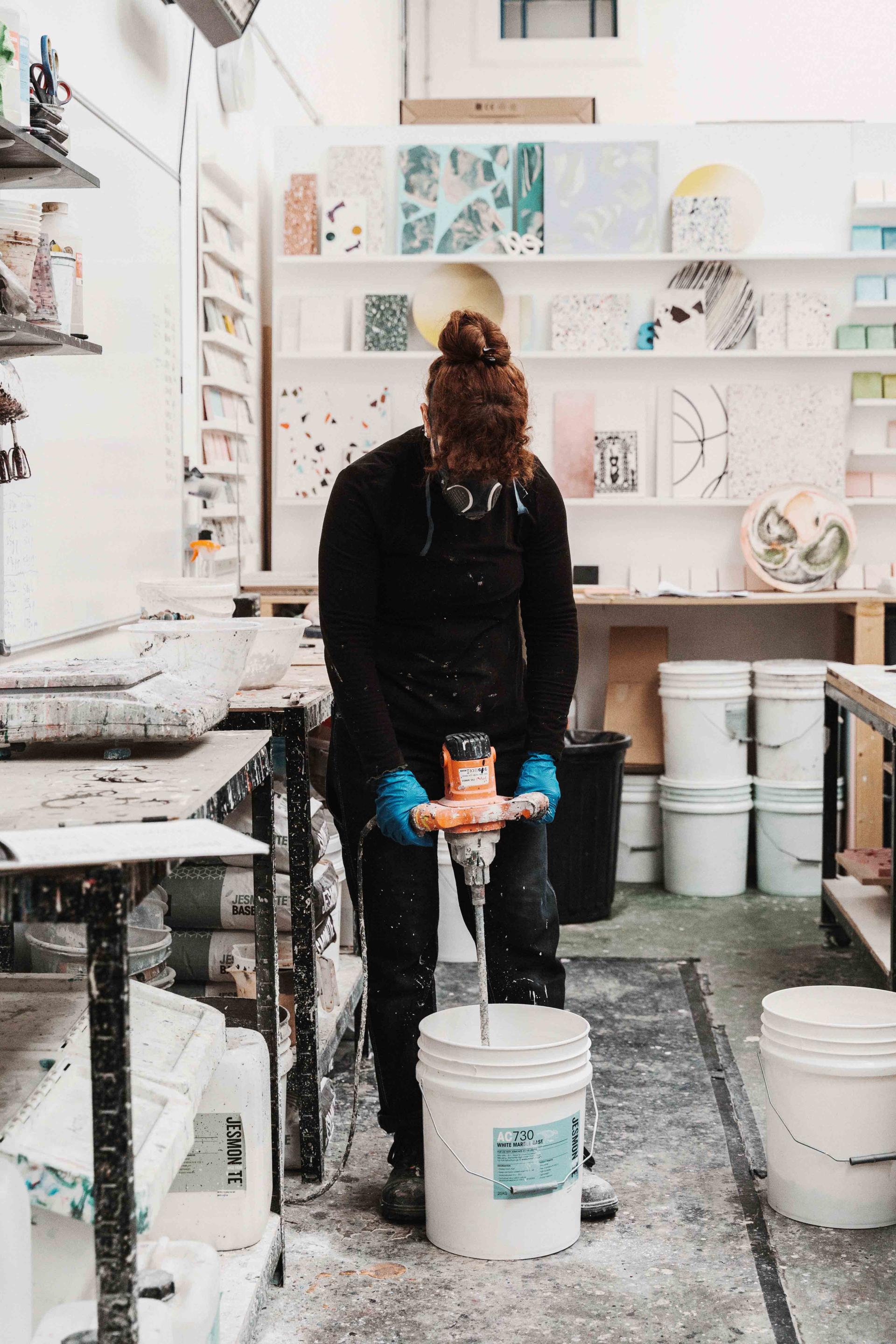 A person mixing ebonite terrazzo materials in a workshop, surrounded by tools and colorful samples on shelves.