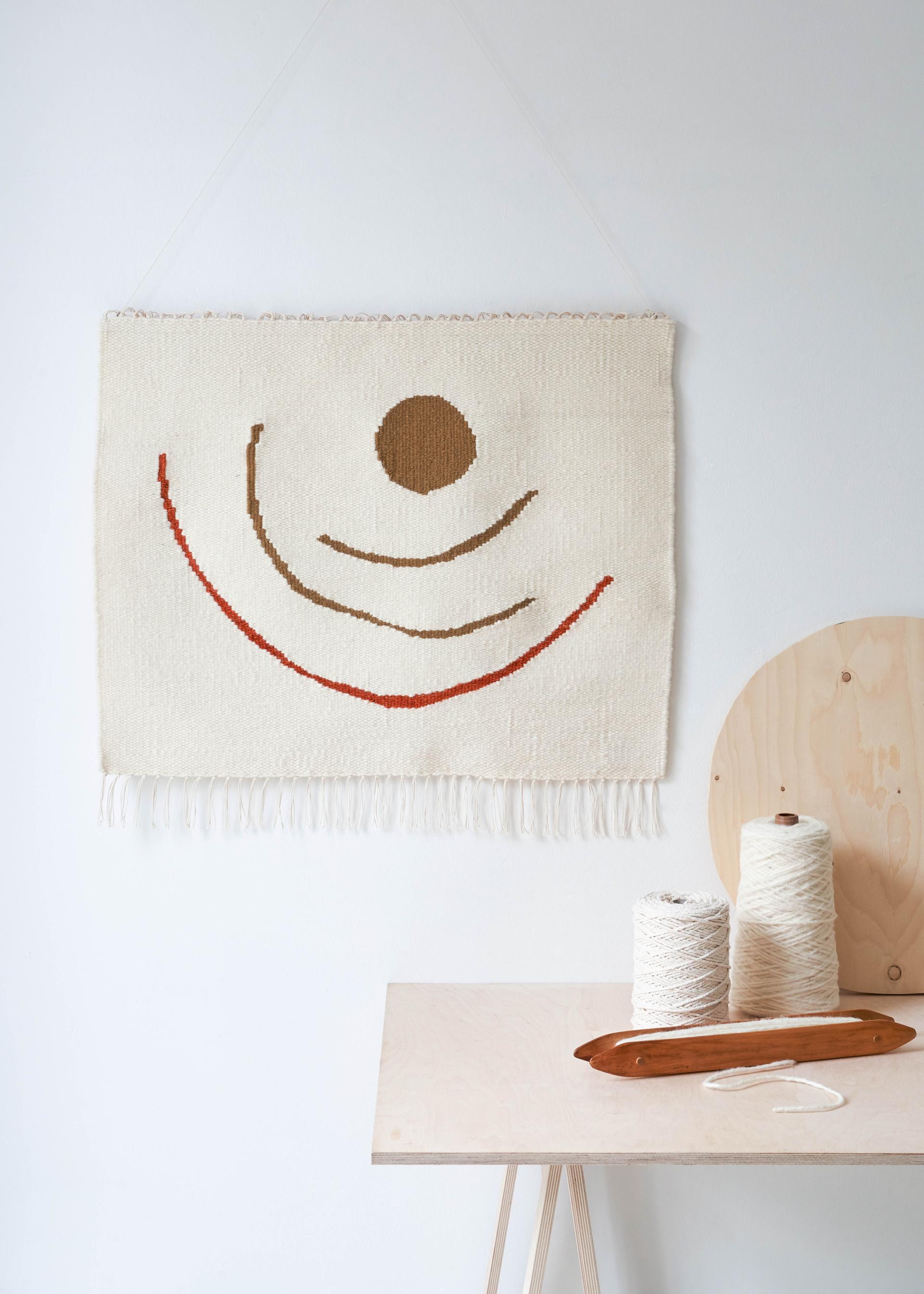 Textile wall hanging featuring abstract patterns, with spools of thread and weaving tools on a wooden table.