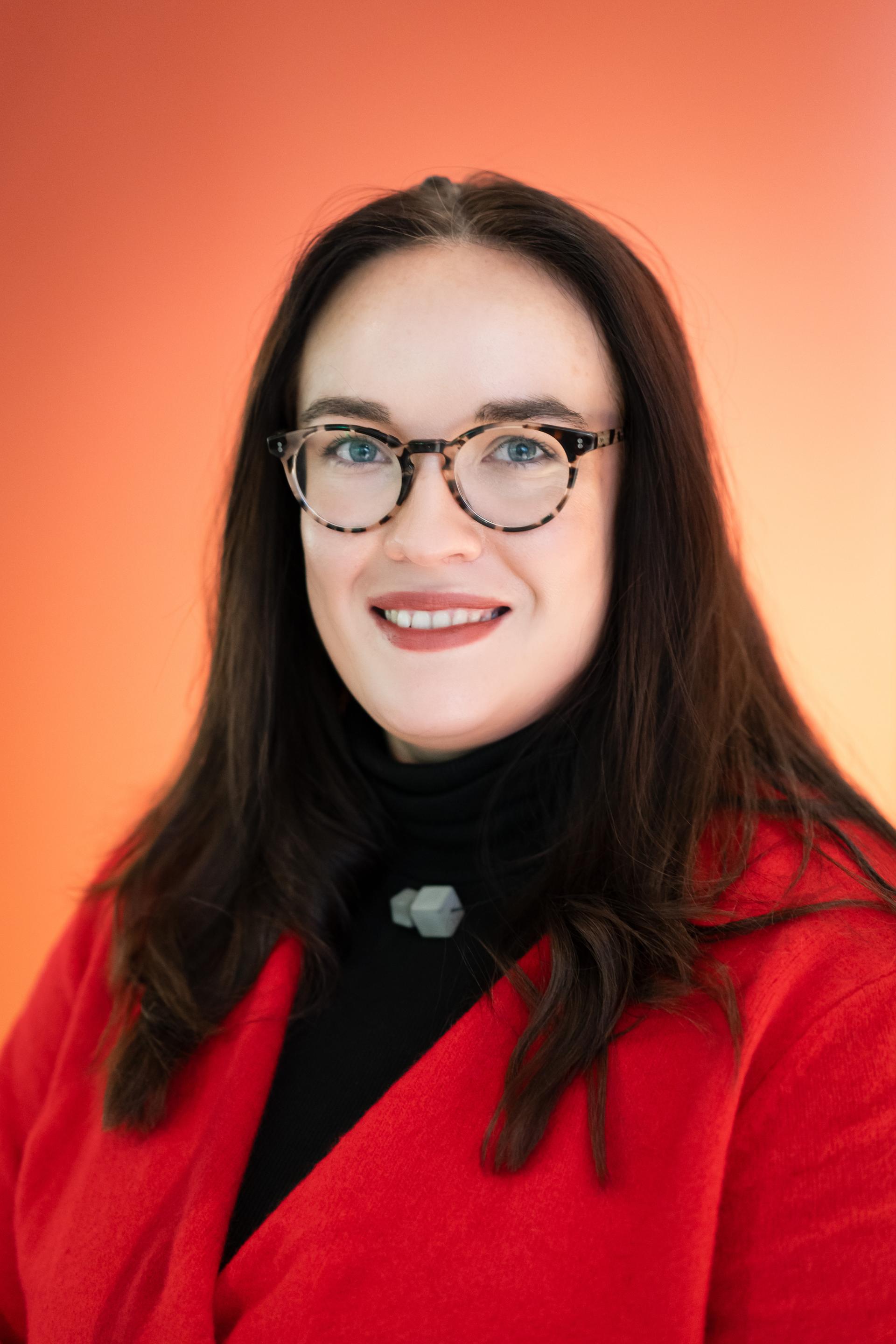 Professional woman with glasses and long dark hair, wearing a red blazer, promoting workplace flexibility and innovation.