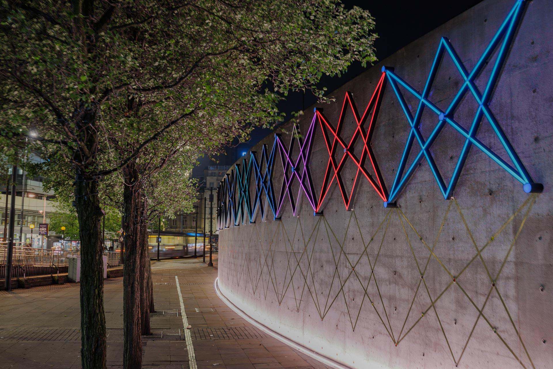 Neon light designs illuminate the wall of Manchester's Piccadilly Gardens Pavilion, surrounded by lush trees at night.