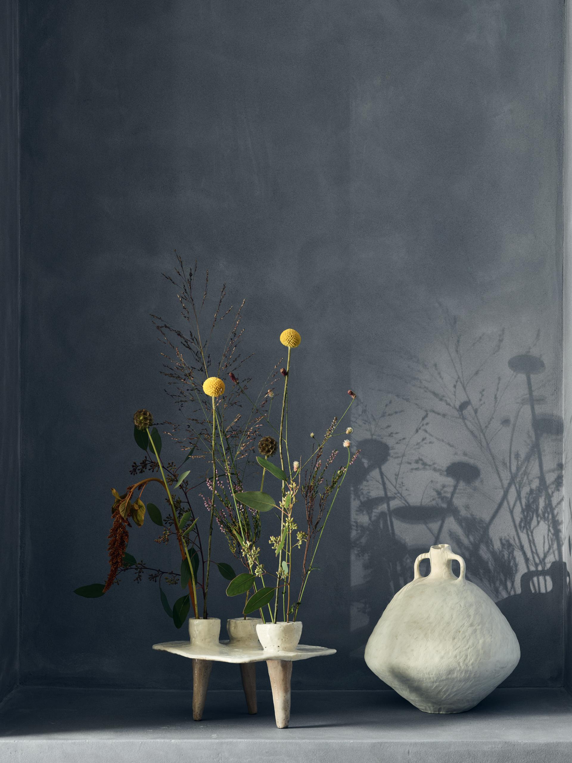 Ceramic vase and handmade flower pots display dried flowers against a textured blue wall, highlighting contemporary craft artistry.