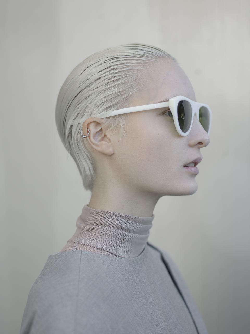 A model with short platinum hair wearing white sunglasses and a stylish outfit, showcasing modern fashion trends.