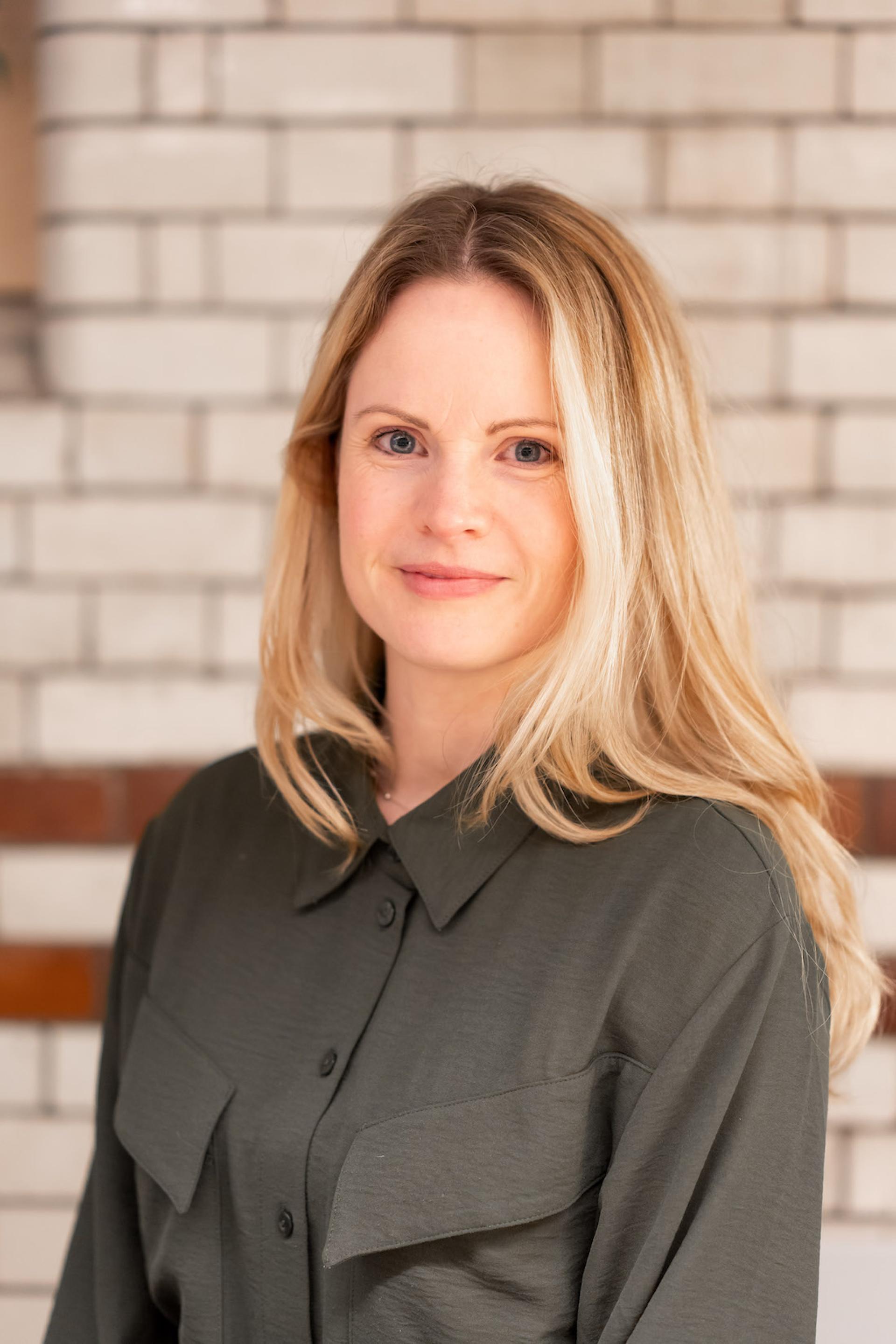 A professional woman with blonde hair, wearing a dark blouse, poses against a textured brick background.