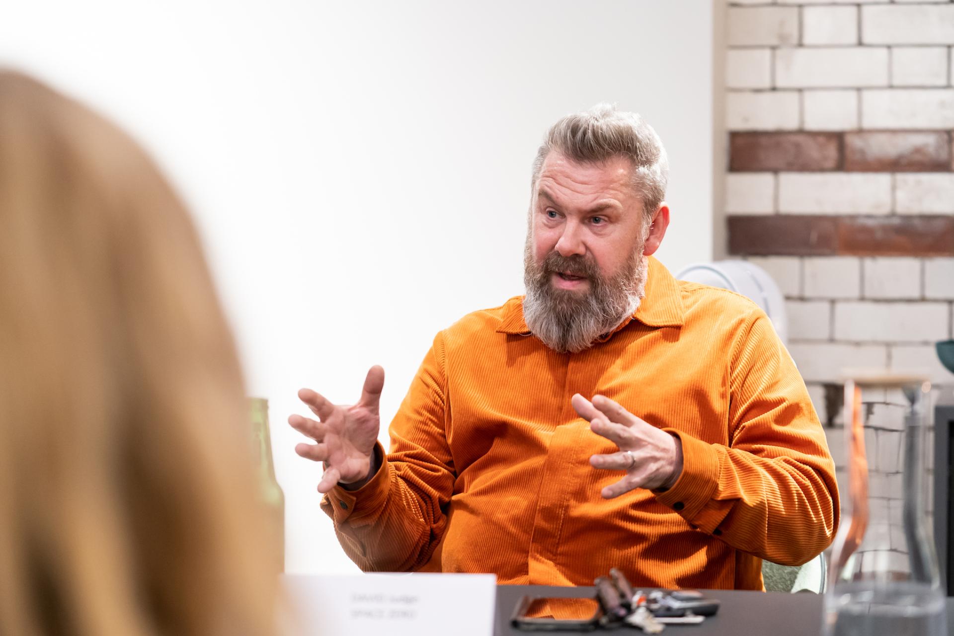A man in an orange shirt passionately discussing the future of education in a modern classroom setting.