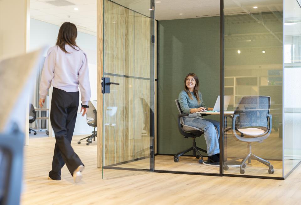 A woman sits at a table in a modern office, using the Vepa Be Hybrid task chair amidst a flexible workspace.