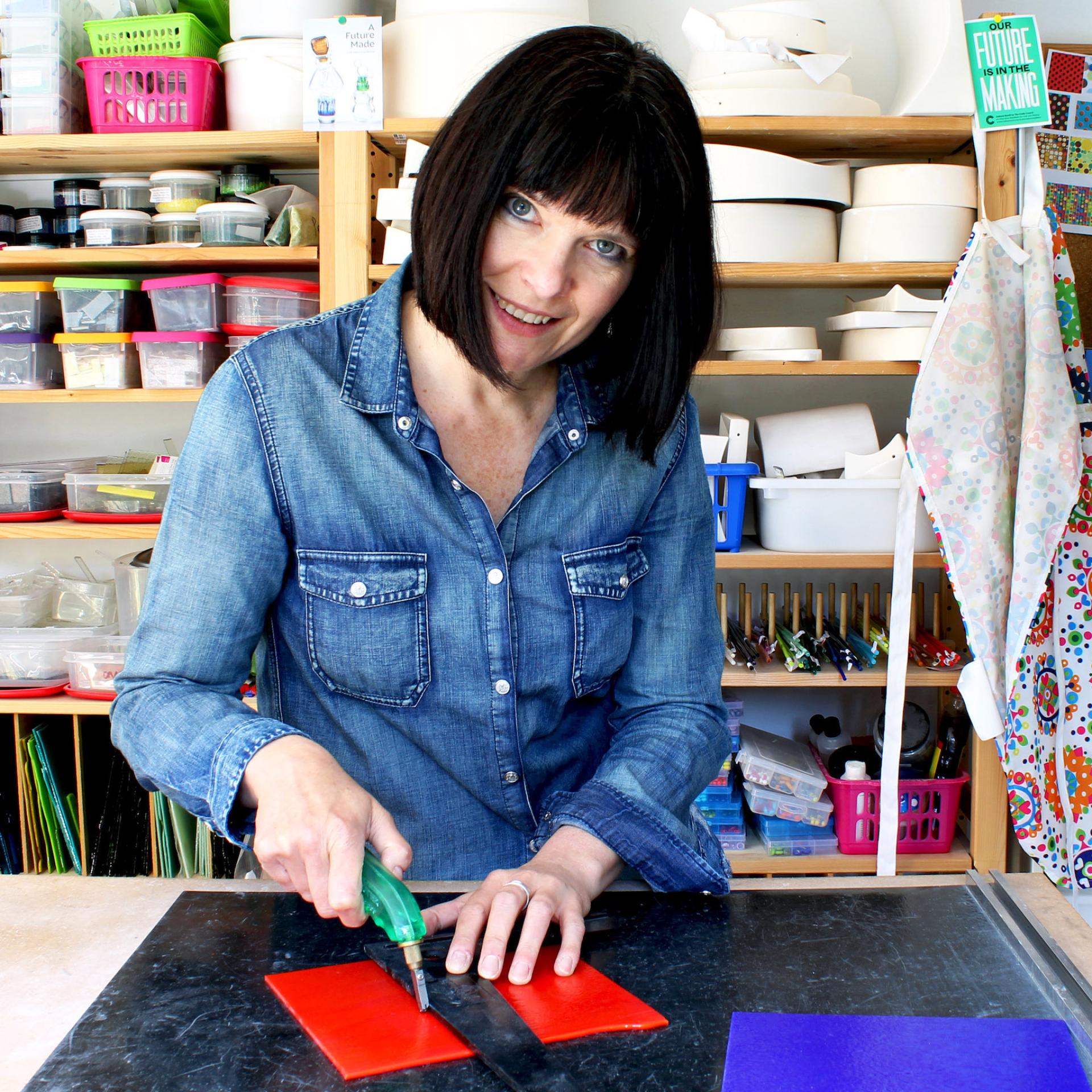 Creative artisan using a cutting tool on red glass pieces in a vibrant craft studio filled with supplies.