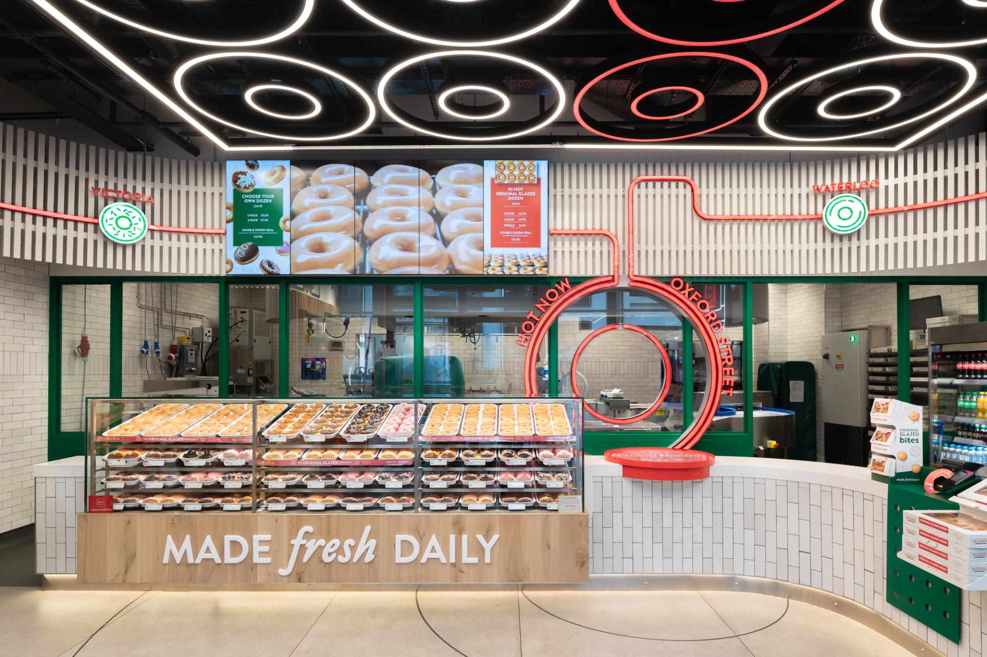Brightly lit Krispy Kreme store interior featuring a display case of freshly made donuts and vibrant neon signage.