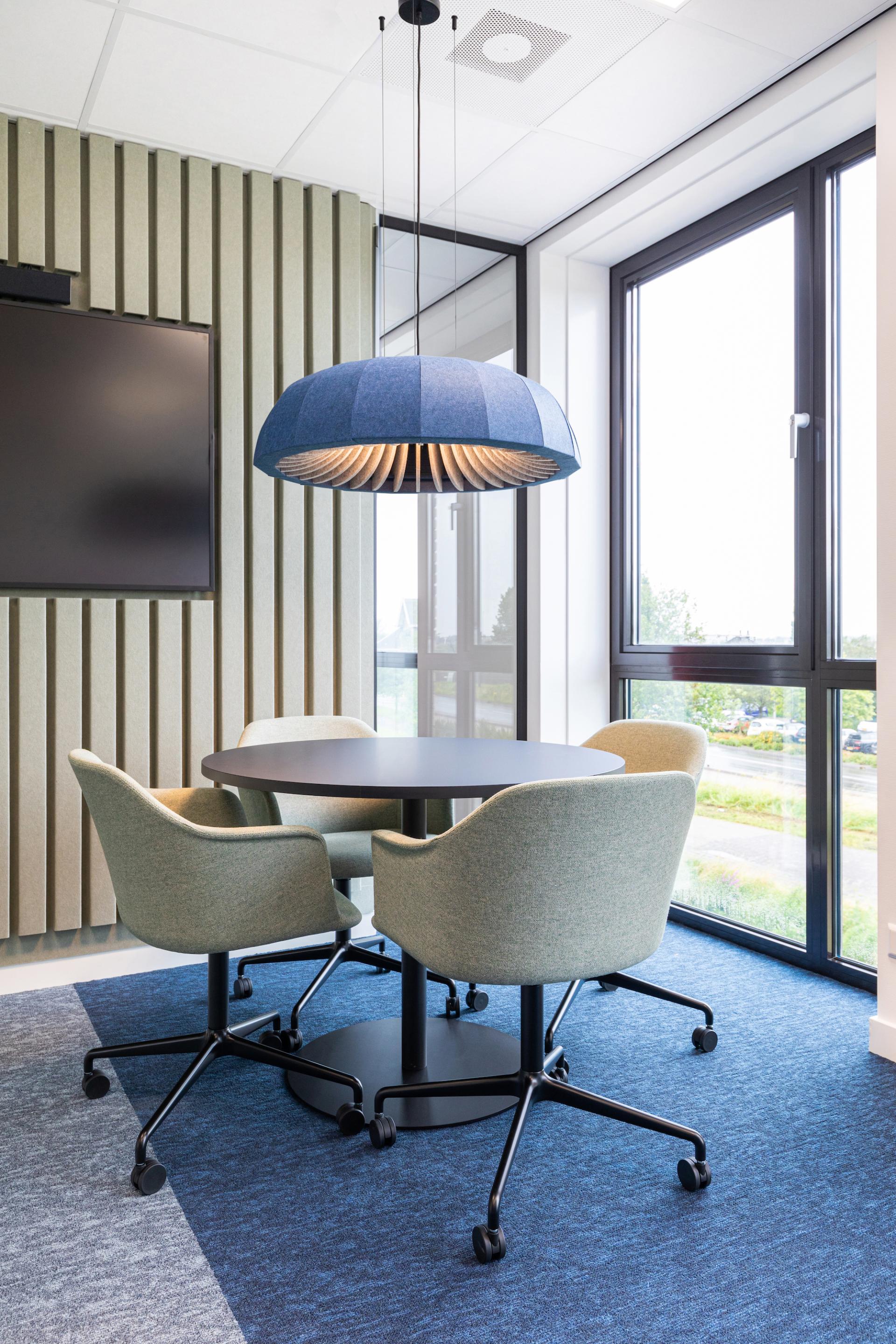 Stylish lumenear acoustic lighting hangs over a modern meeting space with green chairs and large windows, enhancing aesthetics and sound absorption.