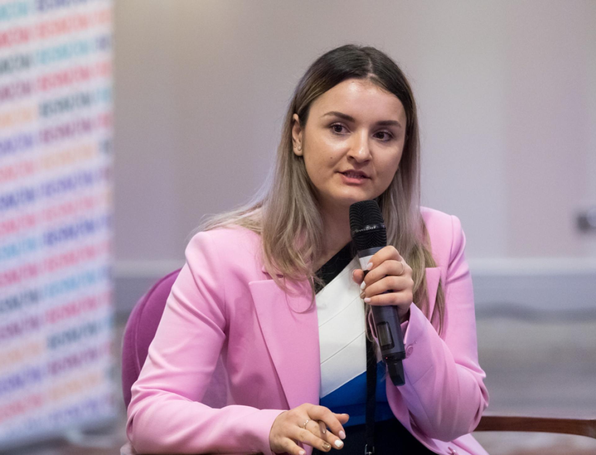 Young woman in a pink blazer speaking into a microphone during an interview event, highlighting diverse perspectives in 2024.