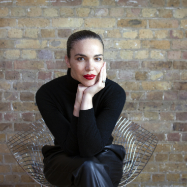 Natasha Bonugli, wearing a black turtleneck, poses thoughtfully in a modern chair against a textured brick wall.