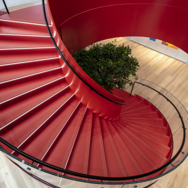 Modern spiral staircase with red steps and glass railing, featuring a plant at the center, in Canopius’ new London office.