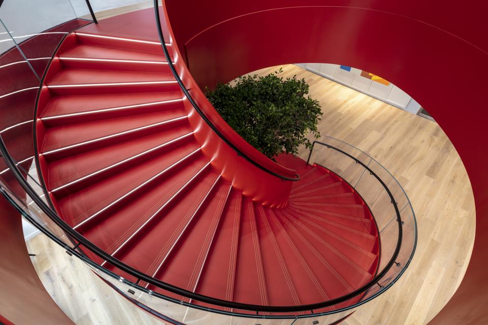 Modern spiral staircase with red steps and glass railing, featuring a plant at the center, in Canopius’ new London office.