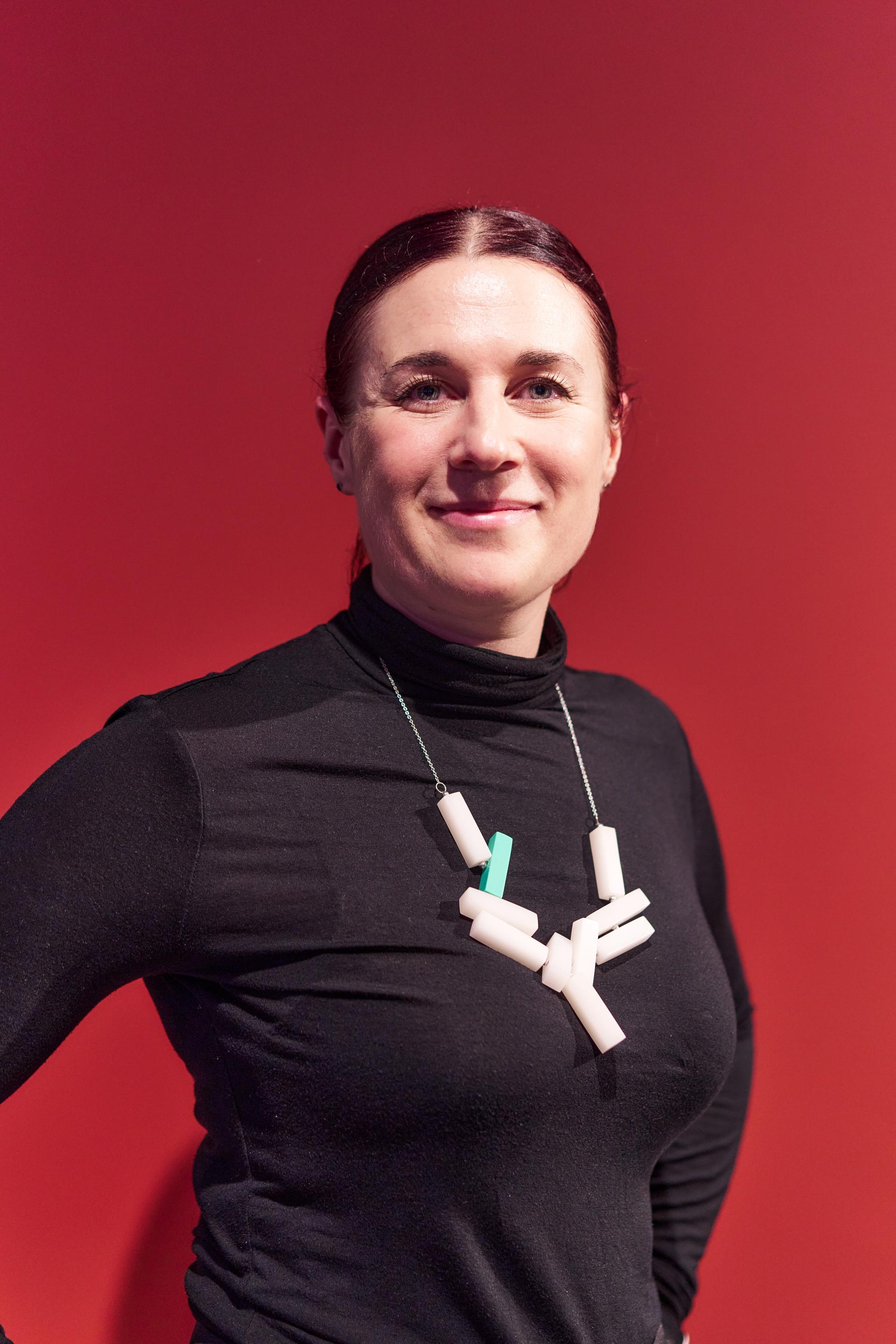 A woman in a black turtleneck with a geometric necklace, posed against a red background, at a Glasgow property event.