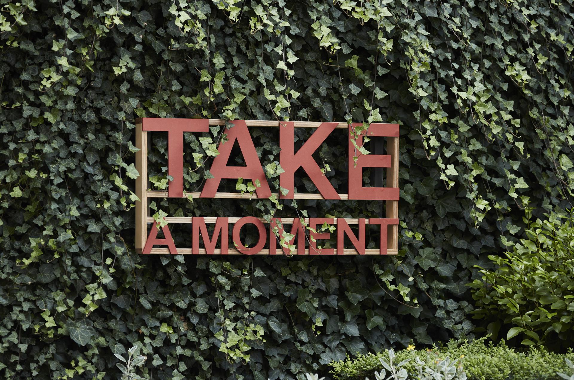 Uplifting sign reading "TAKE A MOMENT," surrounded by lush greenery in Manchester's NOMA neighbourhood.