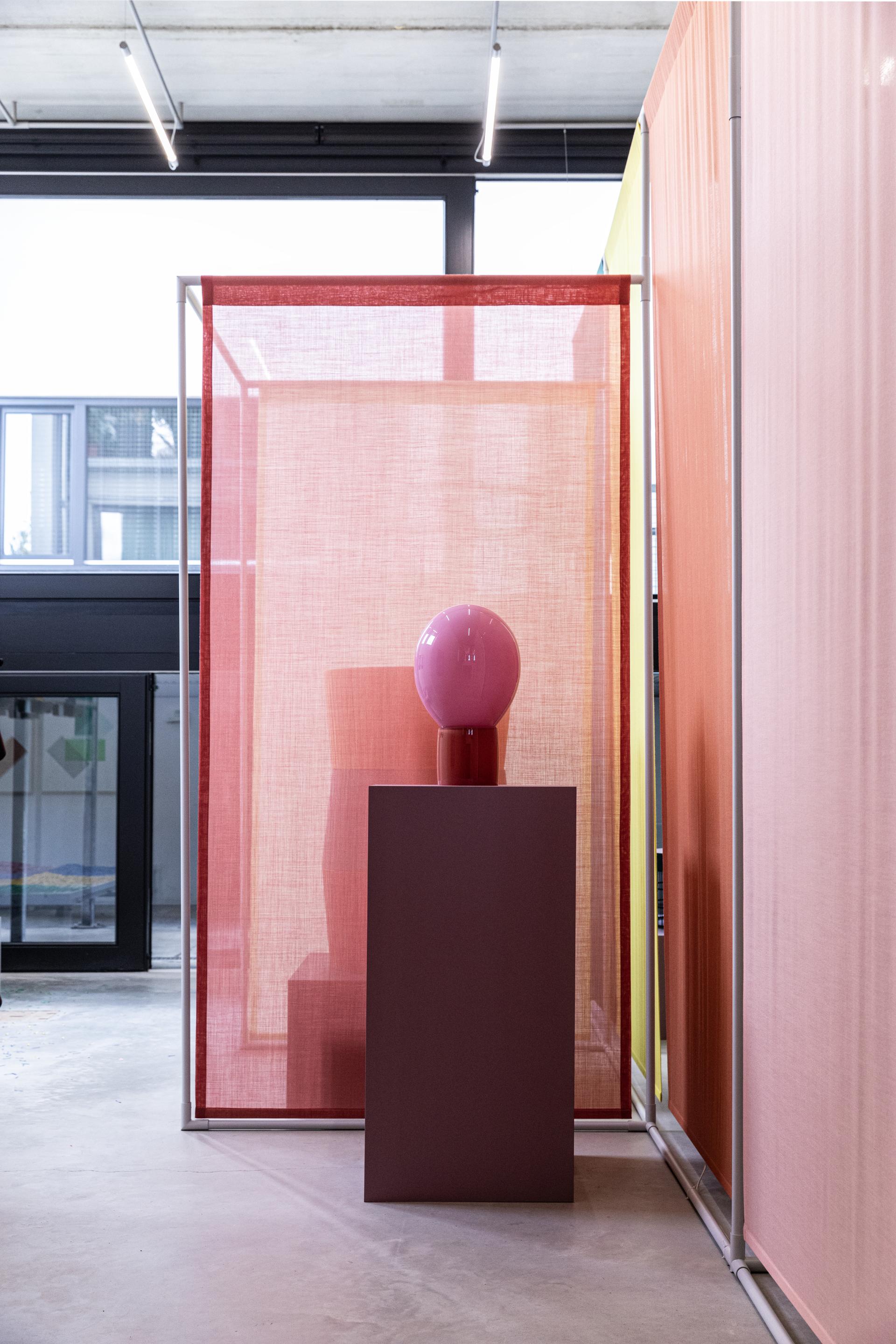 Colorful translucent fabrics frame a pink glass lamp on a brown pedestal, showcasing innovative material design concepts.