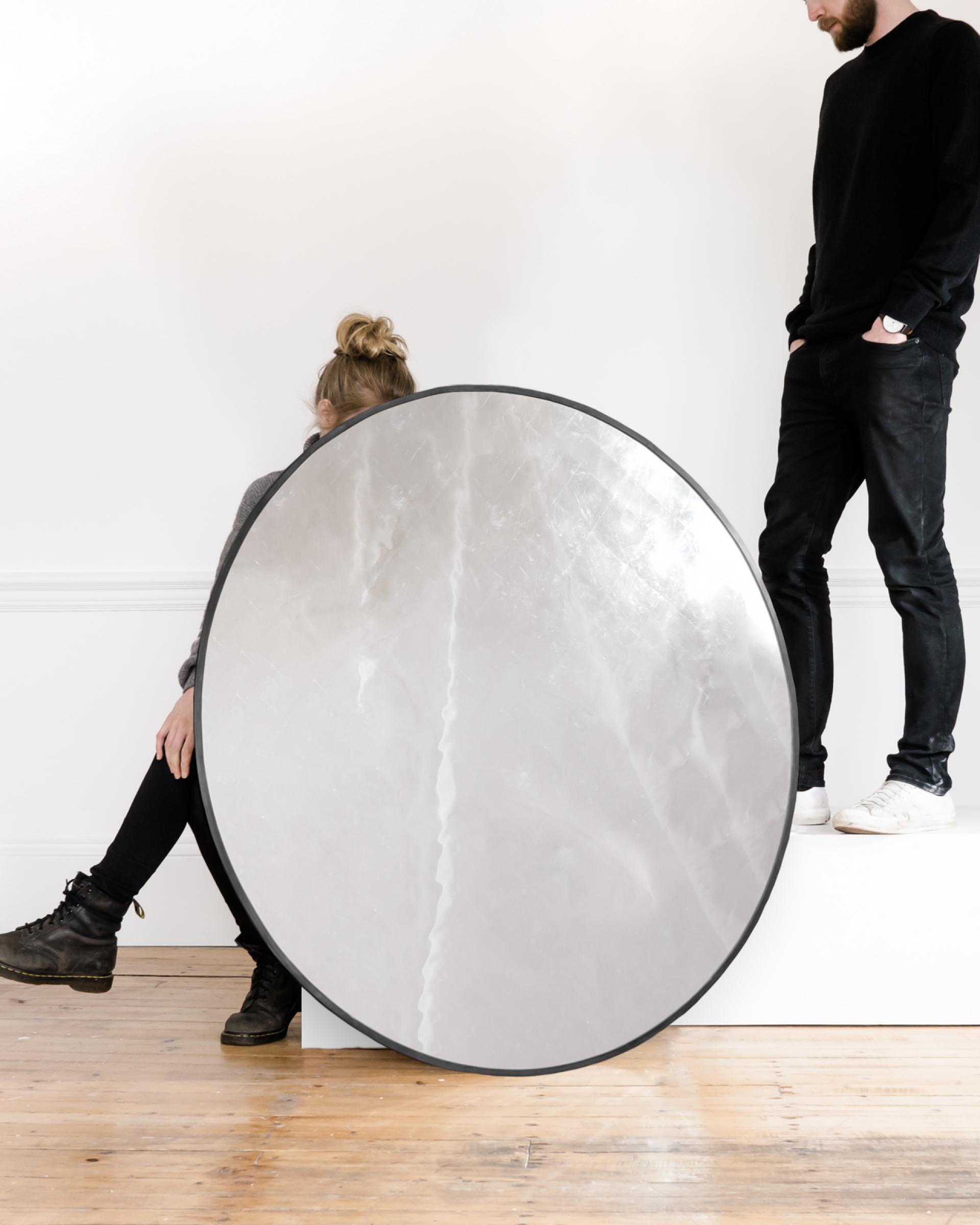 Two individuals pose creatively with a large reflective disc in a bright, minimalist Glasgow studio, showcasing modern craft.
