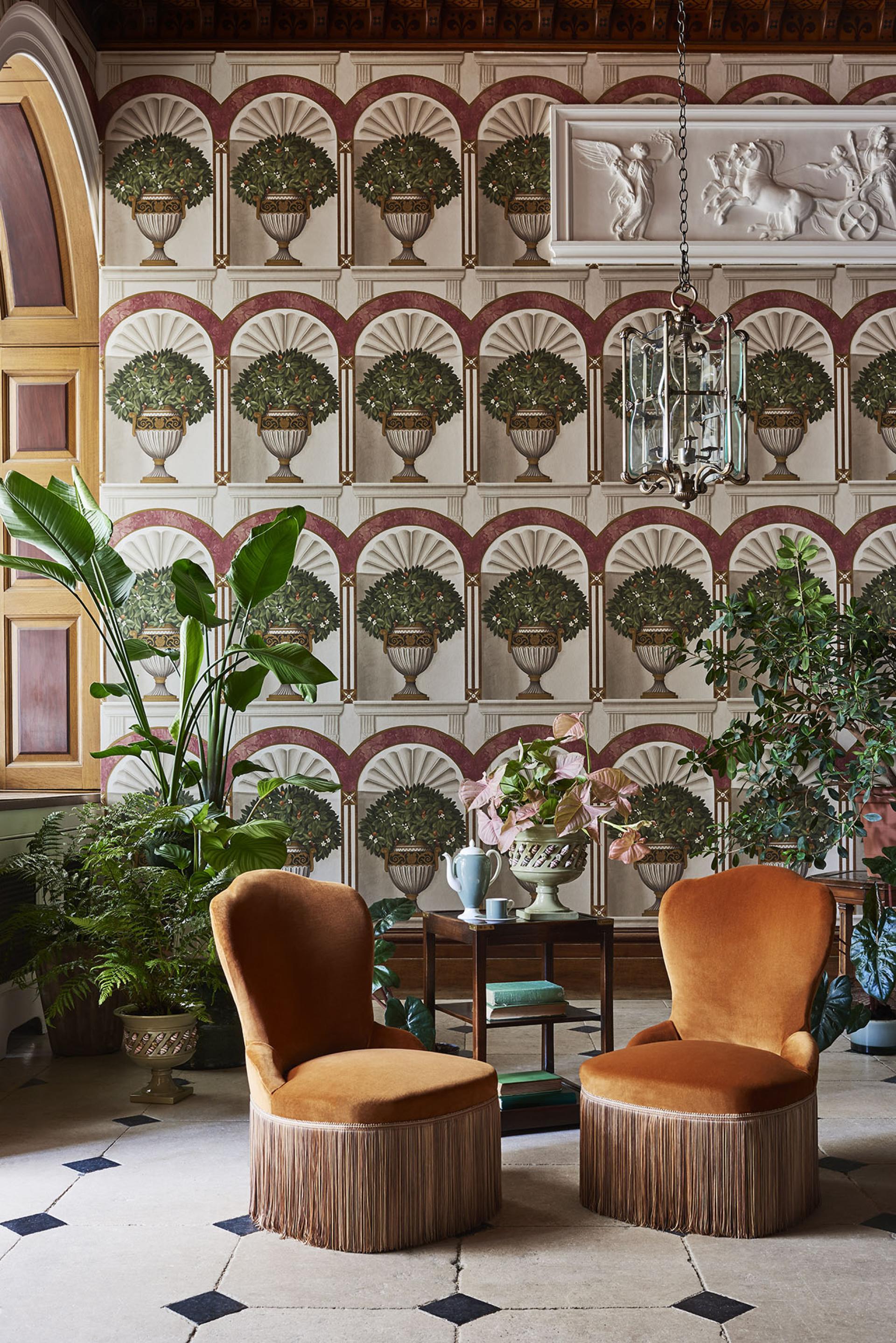 Elegant interior featuring fringed velvet chairs, ornate wallpaper, and lush plants, reflecting British heritage and royal design influences.